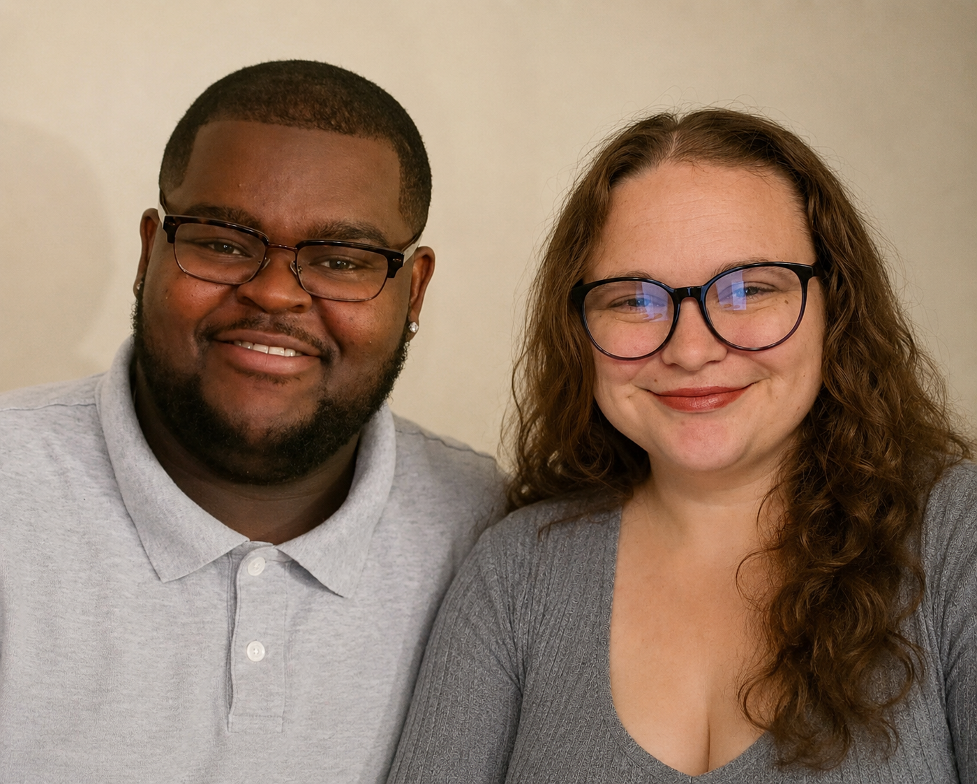 A smiling African American man wearing glasses and a light gray polo shirt standing next to a smiling Caucasian woman with curly brown hair, glasses, and a gray top, against a neutral background.