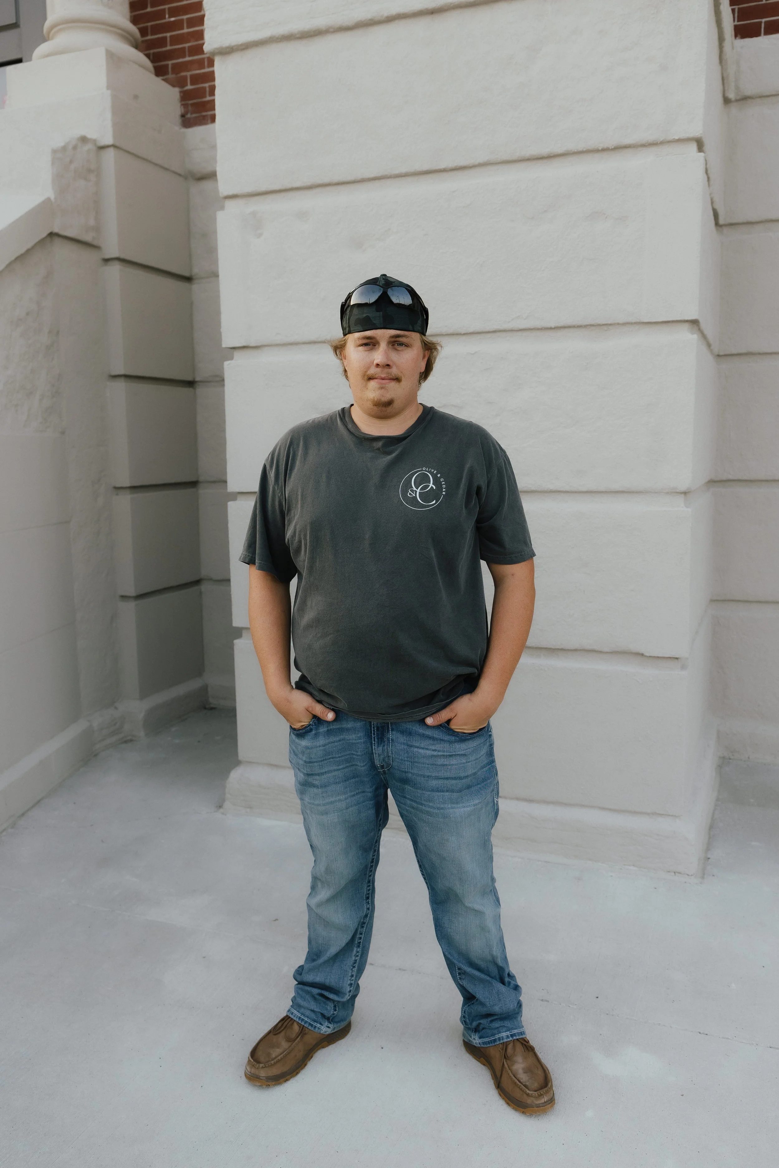 Young man standing outdoors in front of a white stone wall, wearing a black baseball cap with sunglasses on top, a dark T-shirt, blue jeans, and brown work boots.
