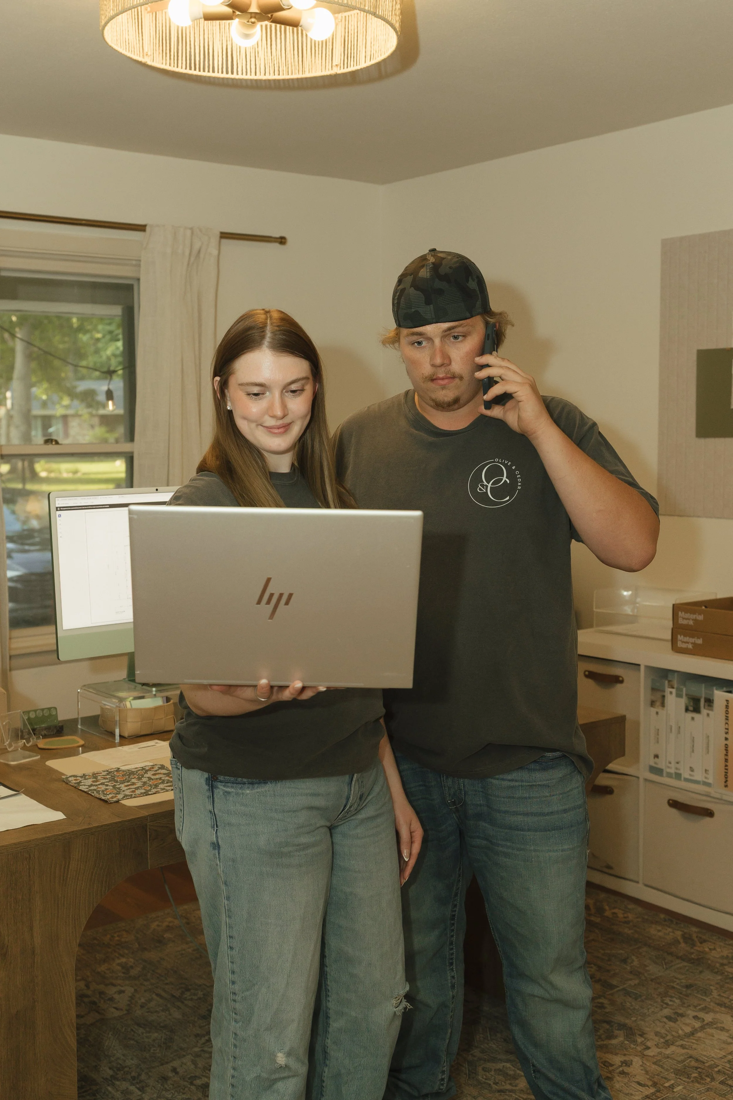 A young woman and a young man are standing together indoors. The young woman is holding a laptop and both are looking at the screen. The man is on the phone and wearing a camouflage cap. The room has a window, a desk with various items, and shelves with binders.