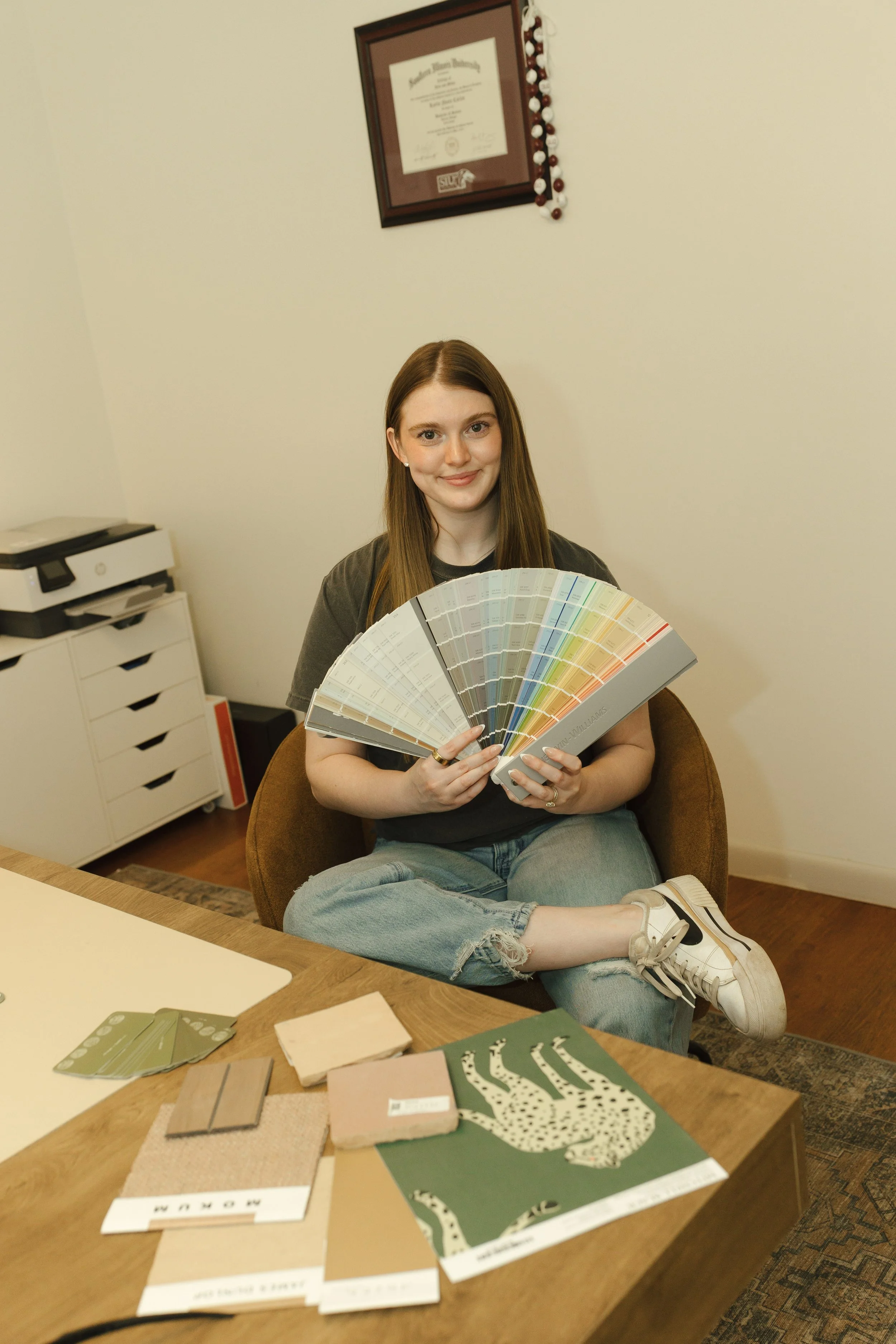 A young woman with long brown hair, wearing a black t-shirt and ripped jeans, sits in a brown chair, smiling, and holding a fan of color sample swatches. In front of her on a wooden table are various design materials, fabric swatches, and mood boards. The background shows an off-white wall with a framed diploma and a beaded garland hanging next to it. A white printer and filing cabinet are also visible.