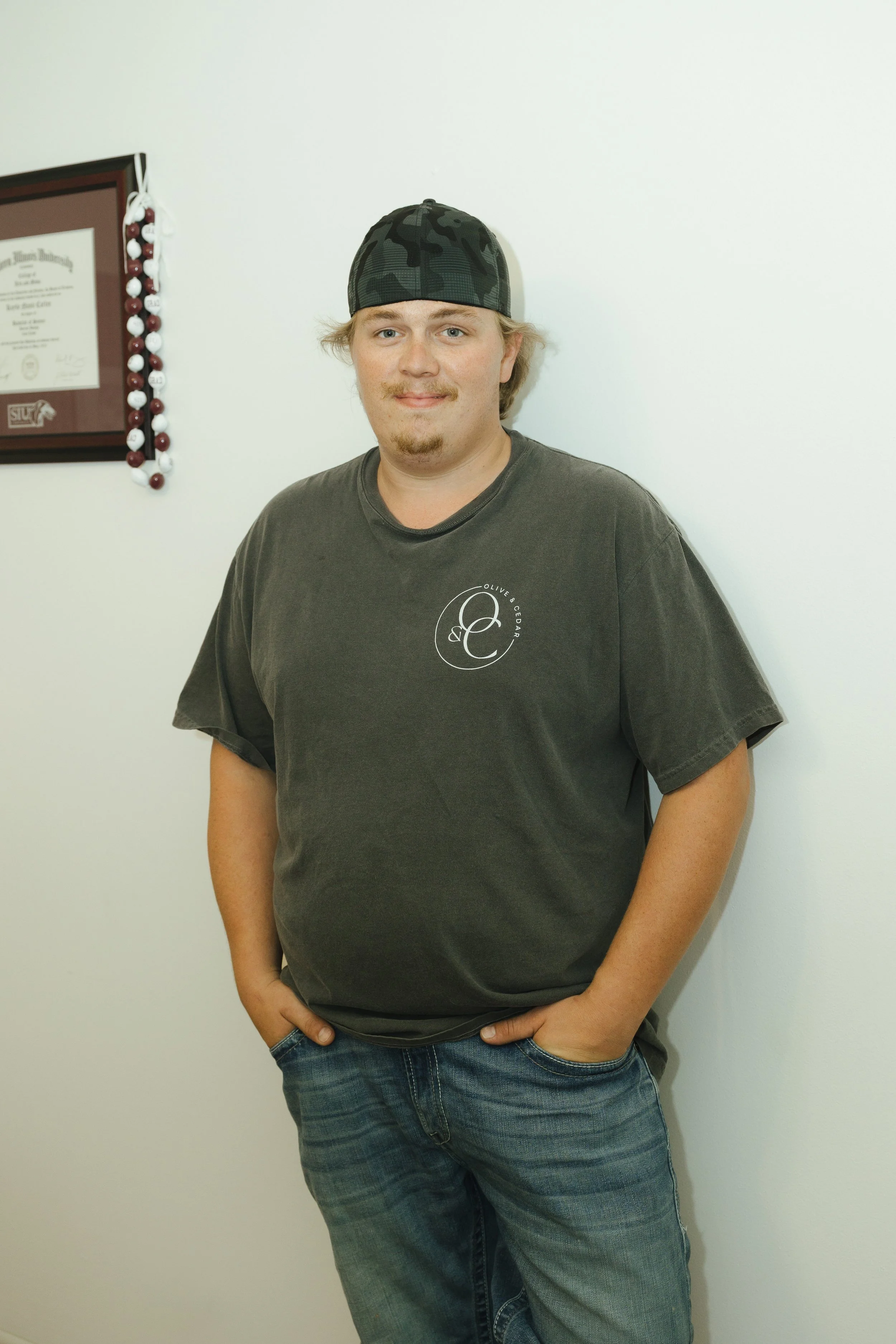 A young man with blonde hair, wearing a black baseball cap and dark t-shirt, stands against a plain white wall with his right hand in his pocket and left hand resting on his hip.