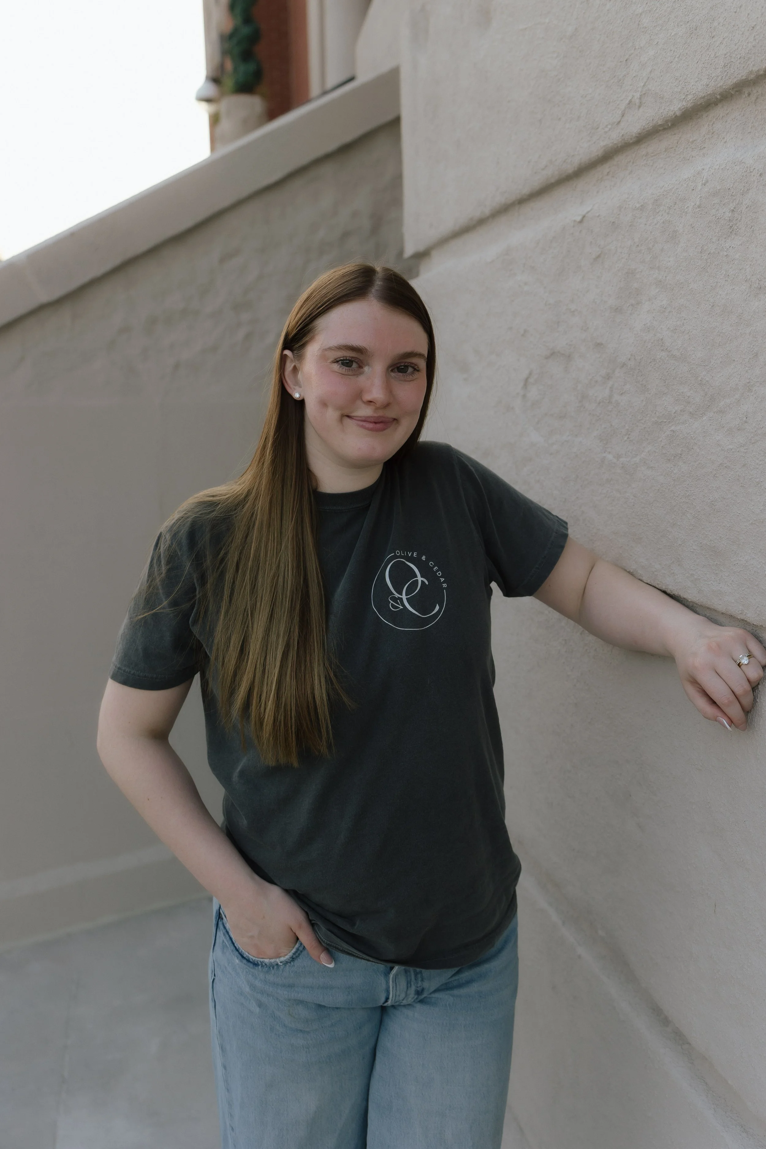 A young woman with long brown hair wearing a black T-shirt and blue jeans, standing outside near a beige concrete wall, smiling at the camera with one hand in her pocket.
