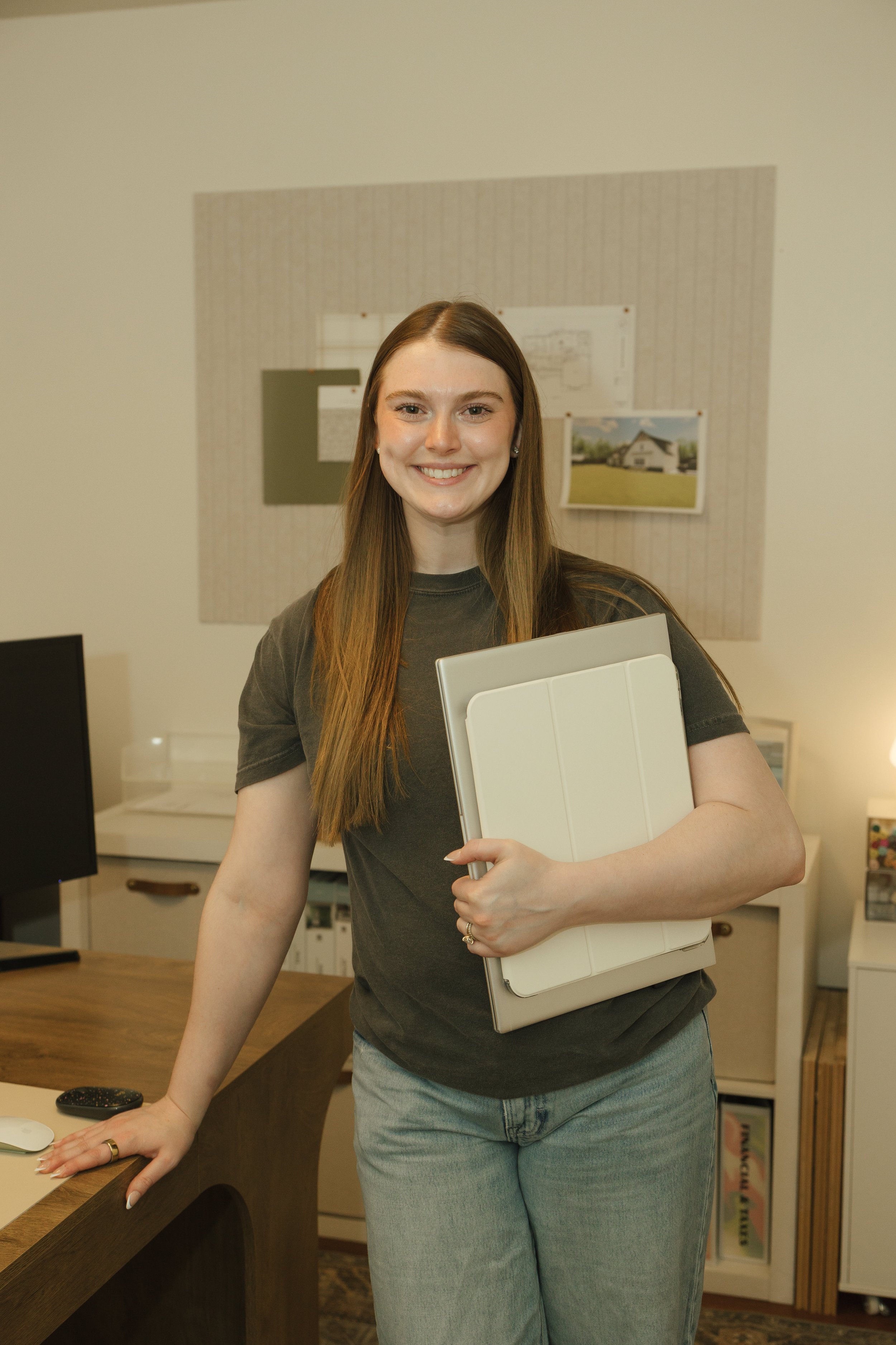 Young woman with long brown hair smiling, holding a closed laptop, standing in an office or home workspace with a desk, computer monitor, and framed pictures on the wall.
