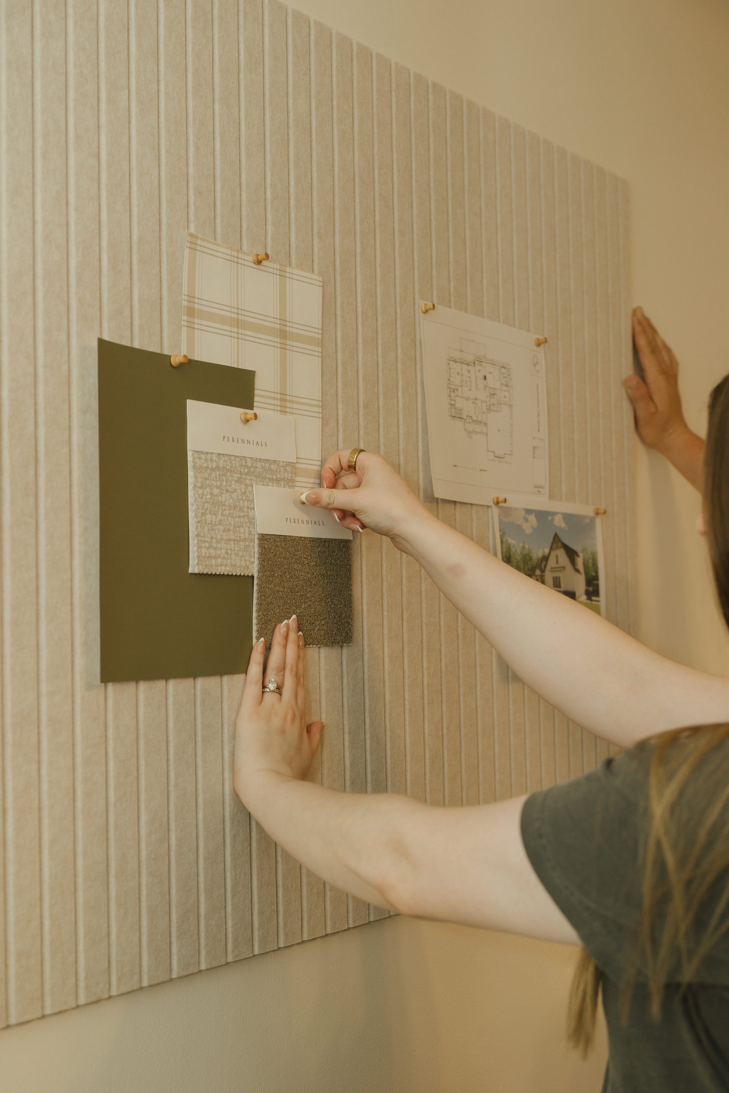 Two people hanging fabric swatches and fabric samples on a bulletin board on a striped wall, with architectural plans and inspiration photos nearby.