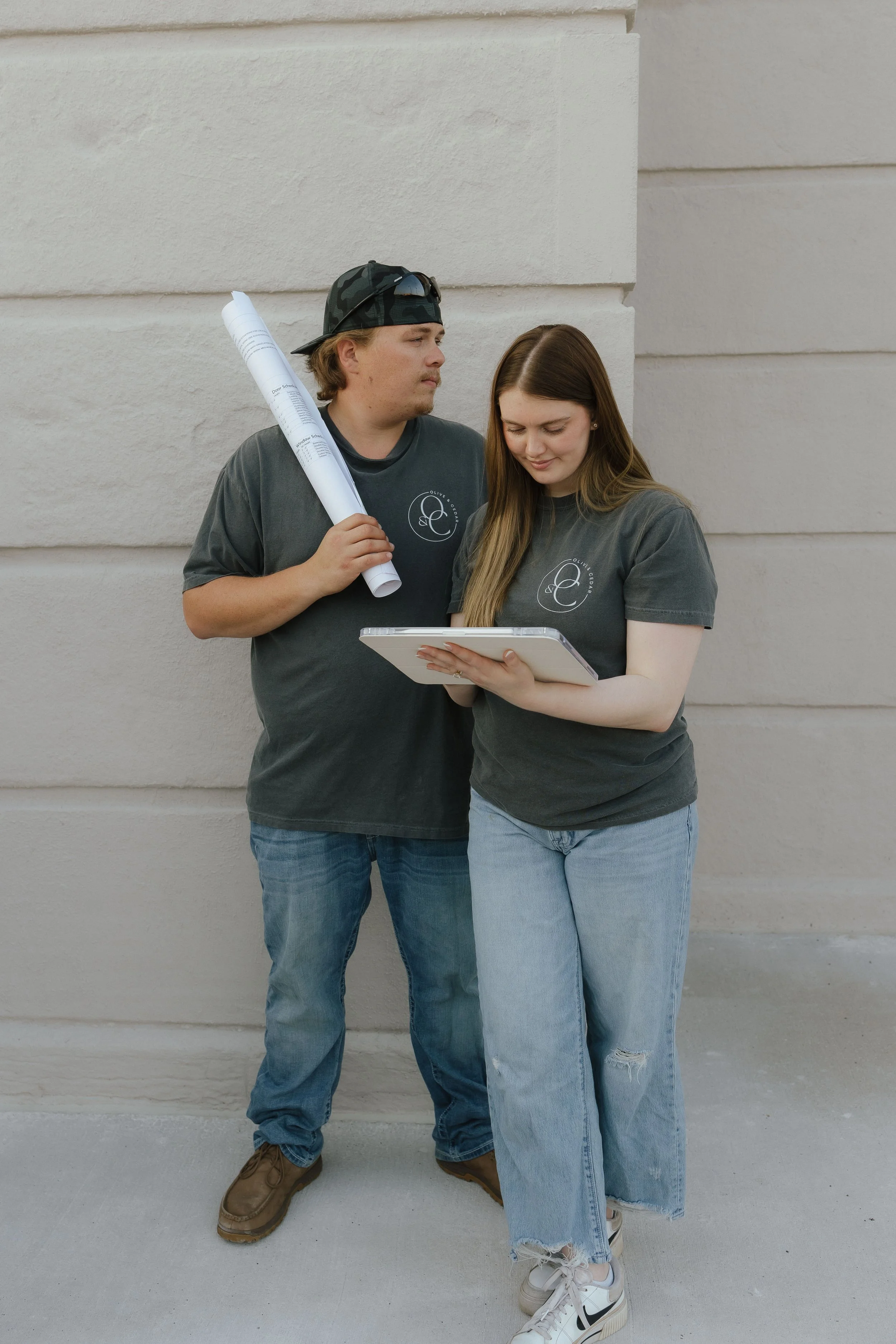 A young man and woman standing together against a white wall. The man is holding a rolled-up document and looking at the woman, who is holding a tablet and looking down at it. They are both wearing casual dark T-shirts with a logo, jeans, and sneakers.