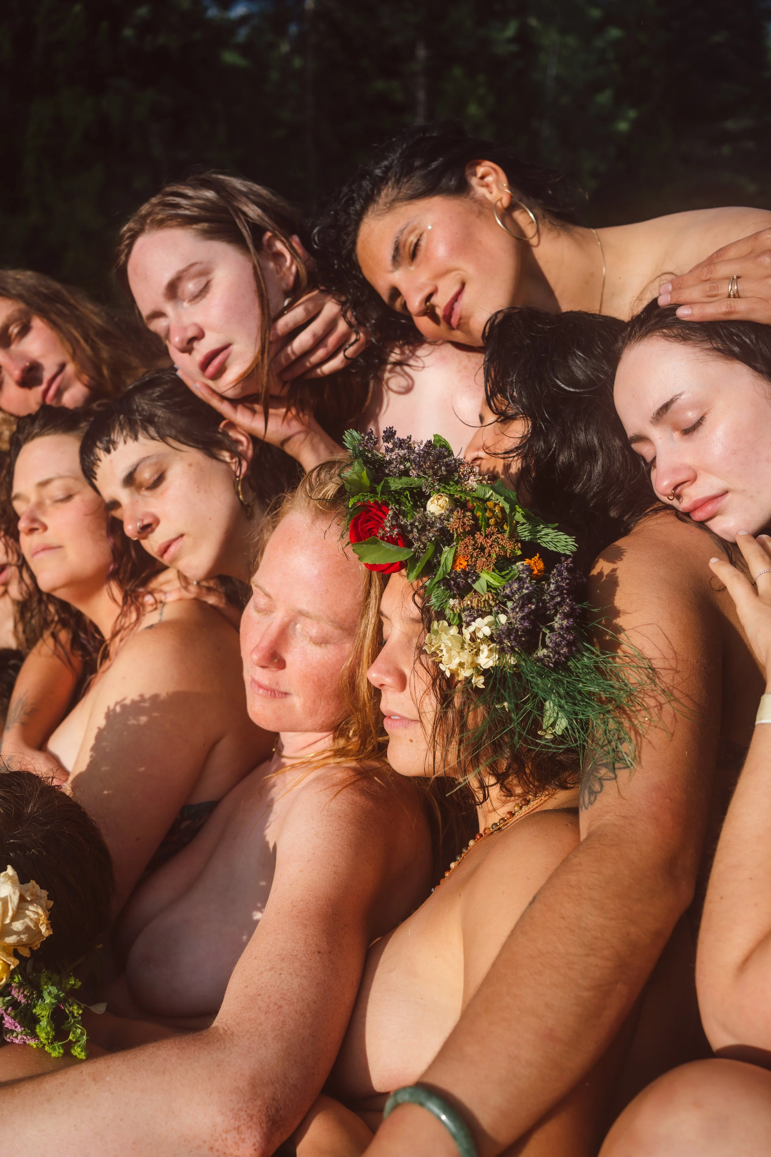 A group of women with closed eyes, some with flowers in their hair, are closely pressed together and seem to be participating in a ritual or ceremony outdoors.