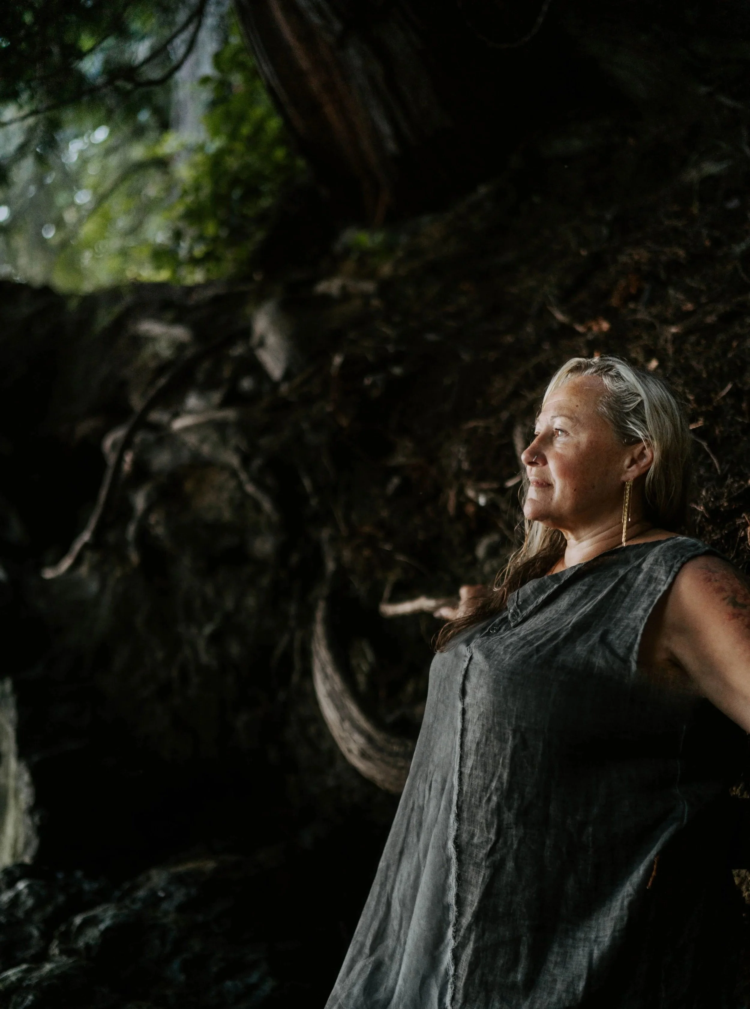 A woman with long gray hair and earrings stands in a dark, earthy cave or forest setting, looking to her right with a slight smile.