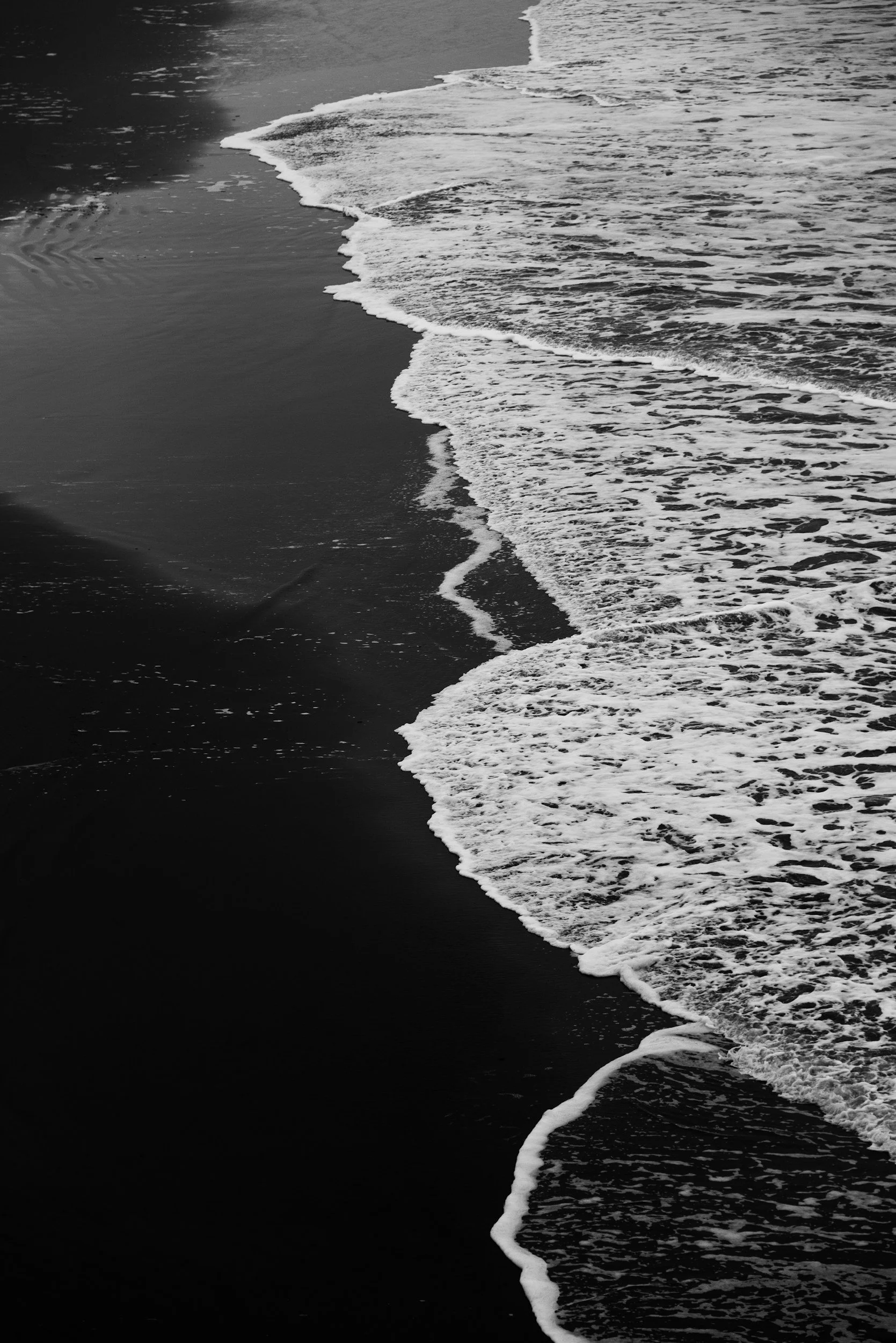 Black and white photo of ocean waves on a sandy beach with foam outlining the shoreline.