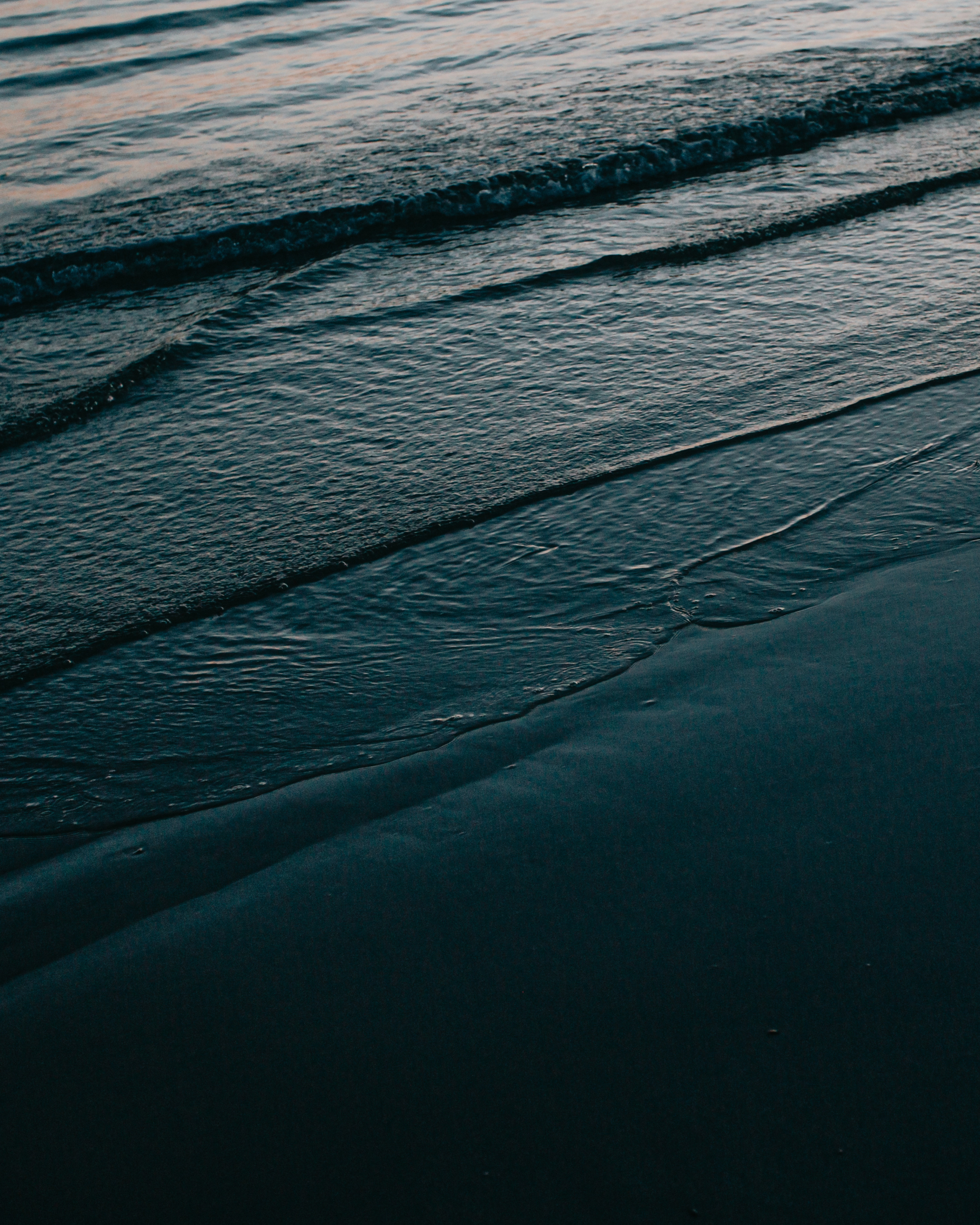Close-up of wet black sand with small waves gently lapping at the shoreline during sunset.