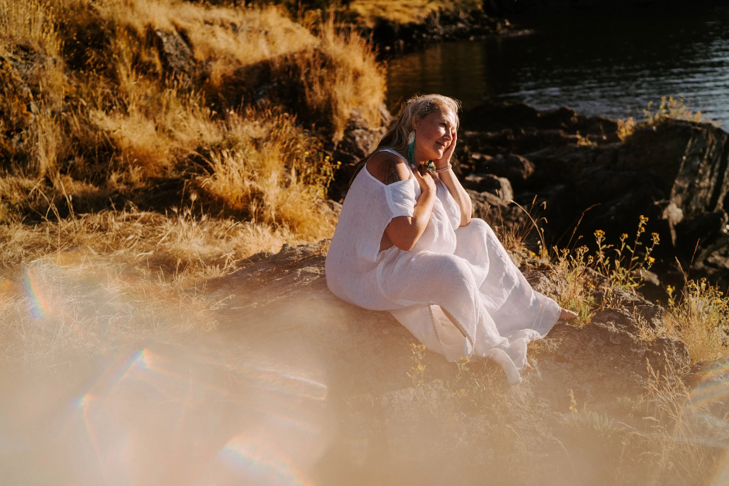 A woman with long blonde hair sitting on rocks by a body of water, wearing a white dress and earrings, smiling and touching her face, during sunset with warm lighting.