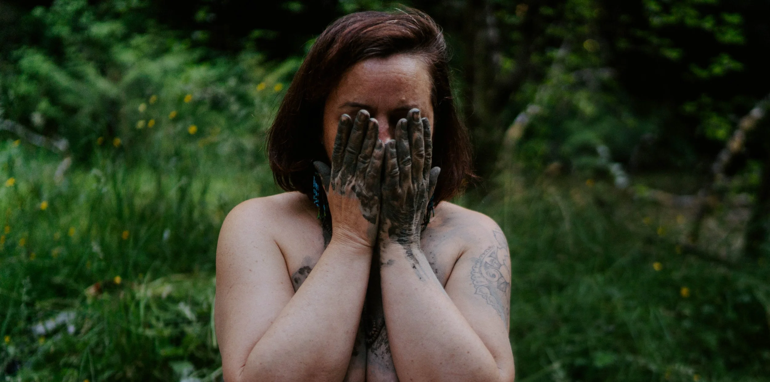 A woman with brown hair and tattoos covering her arms and chest, holding her face with both hands, standing outdoors with greenery in the background.