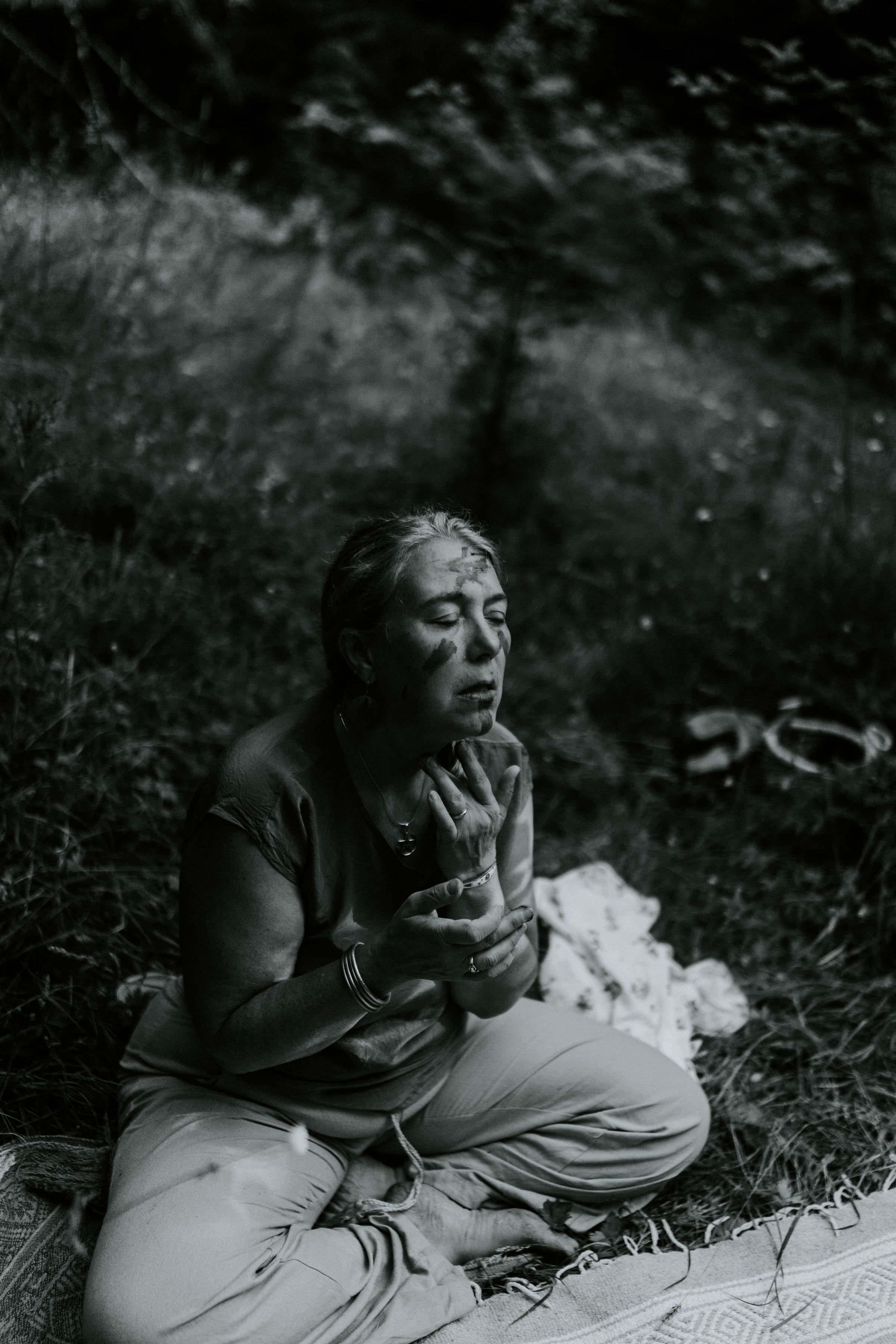 A woman sitting cross-legged outdoors on a mat, practicing meditation with eyes closed and hands in a prayer position, surrounded by natural scenery.