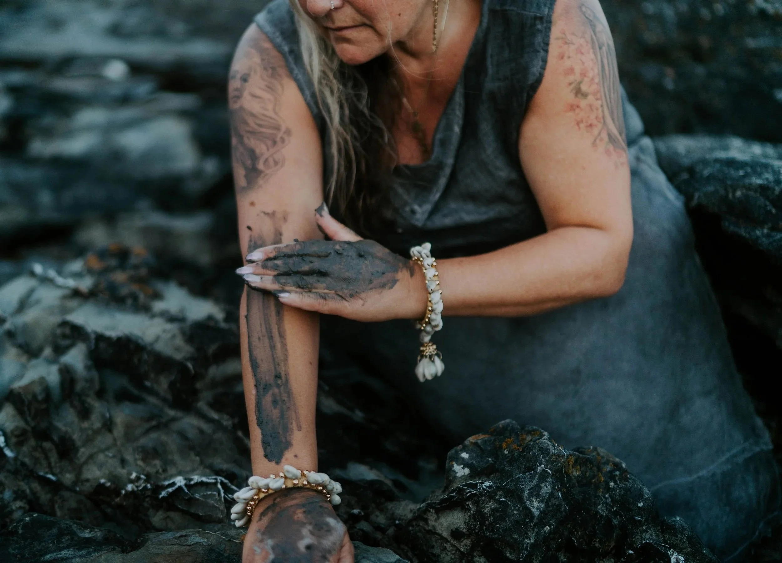 A woman with long blonde hair, tattoos on her arm, and jewelry is sitting on black rocks, possibly near a river or shoreline, with her hand resting on the rocks.