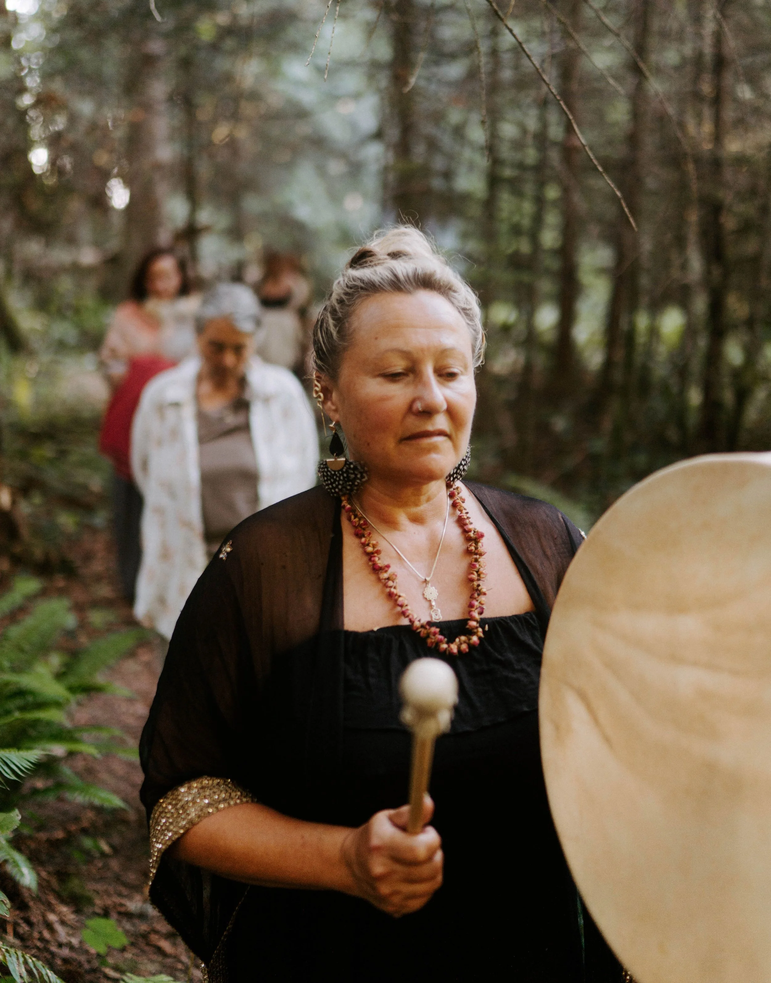 A woman with blonde hair styled in a bun, wearing jewelry and a black top, holding a round wooden shield and a stick, participating in a cultural or medieval reenactment outdoors in a forest.