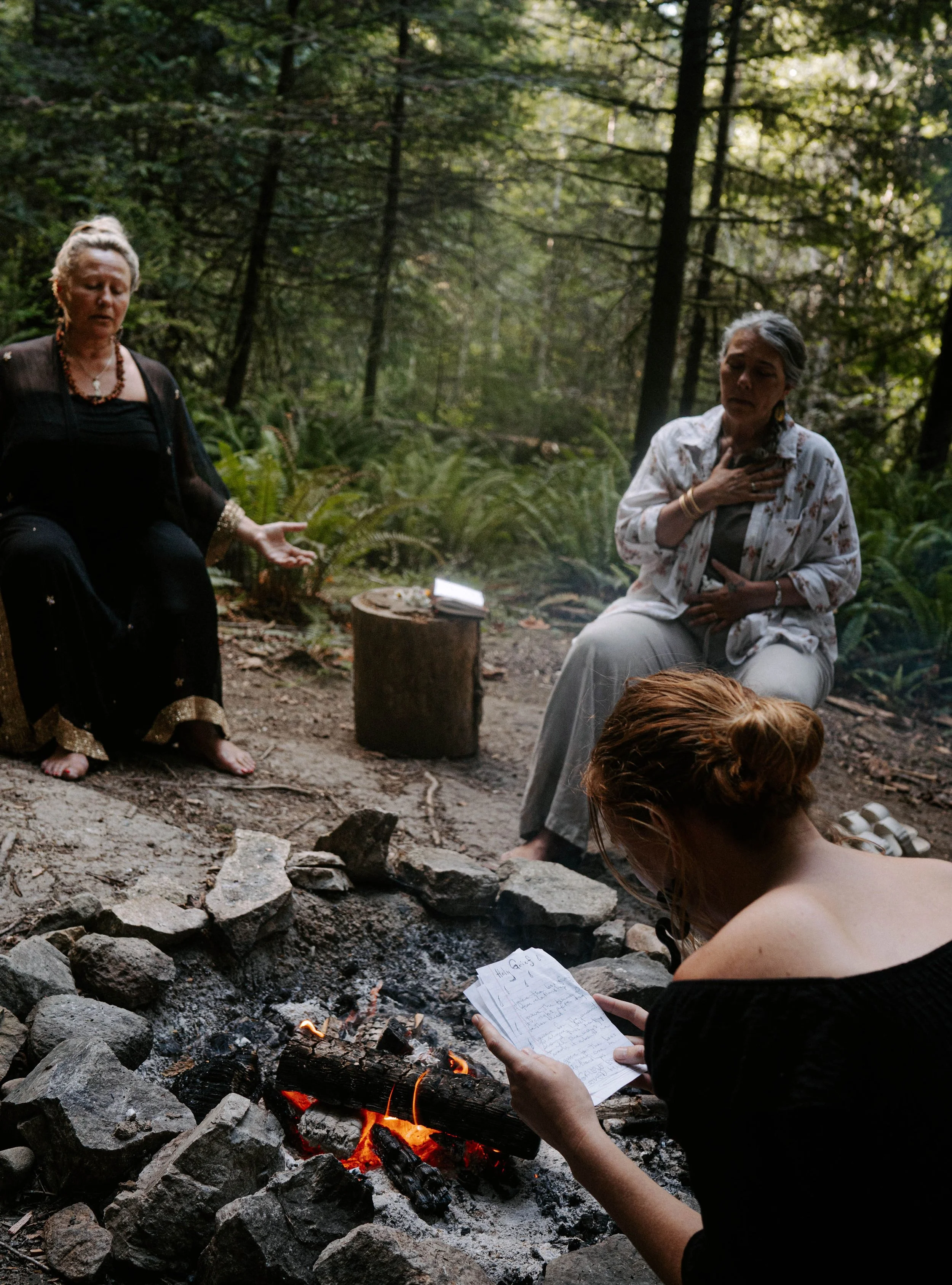 Two women meditate near a campfire in a forest, one sitting with her eyes closed and another reading a handwritten note.