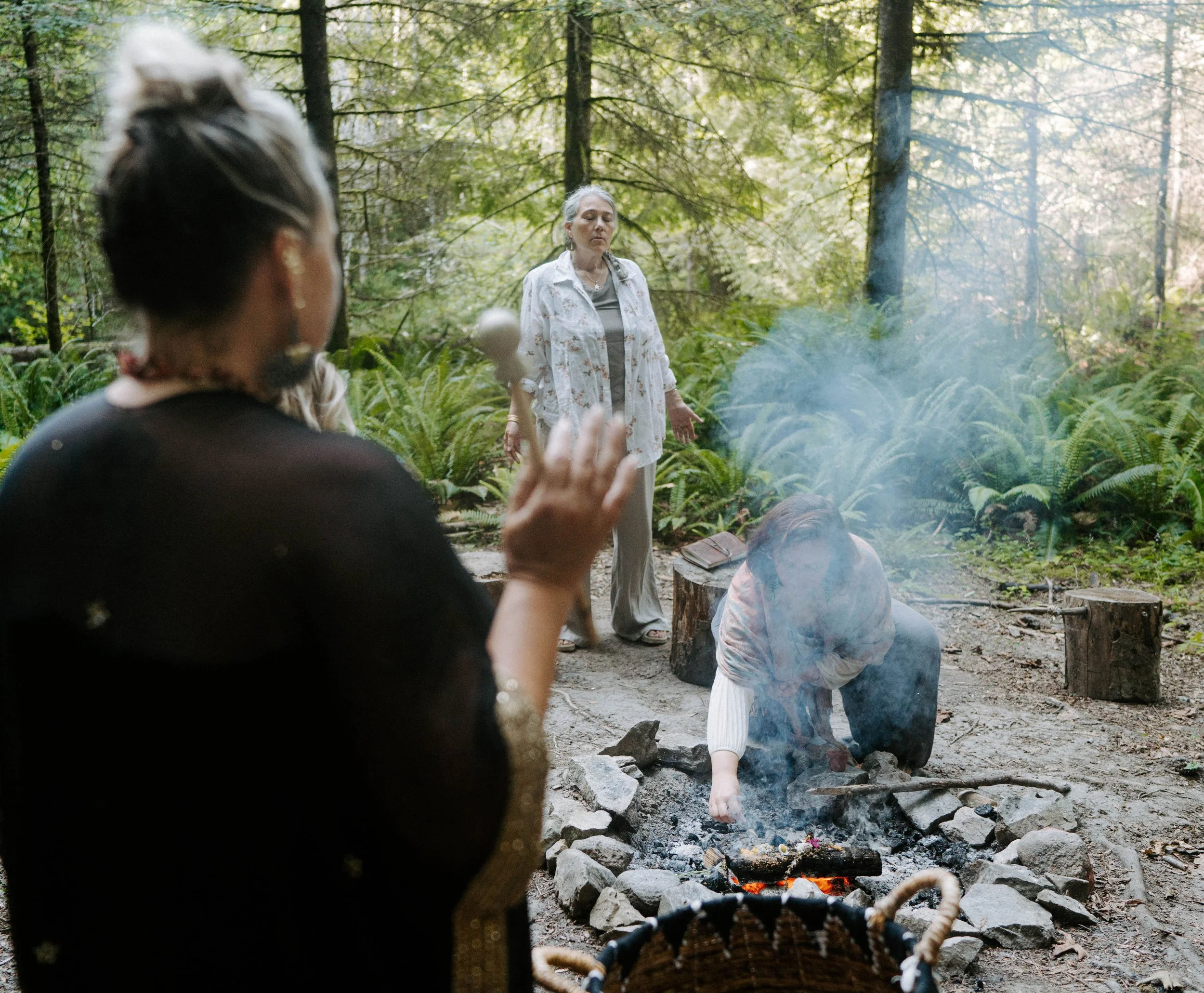 Two women in a forest setting near a campfire, one with gray hair standing with eyes closed and the other with dark hair in the foreground, partially blurred.