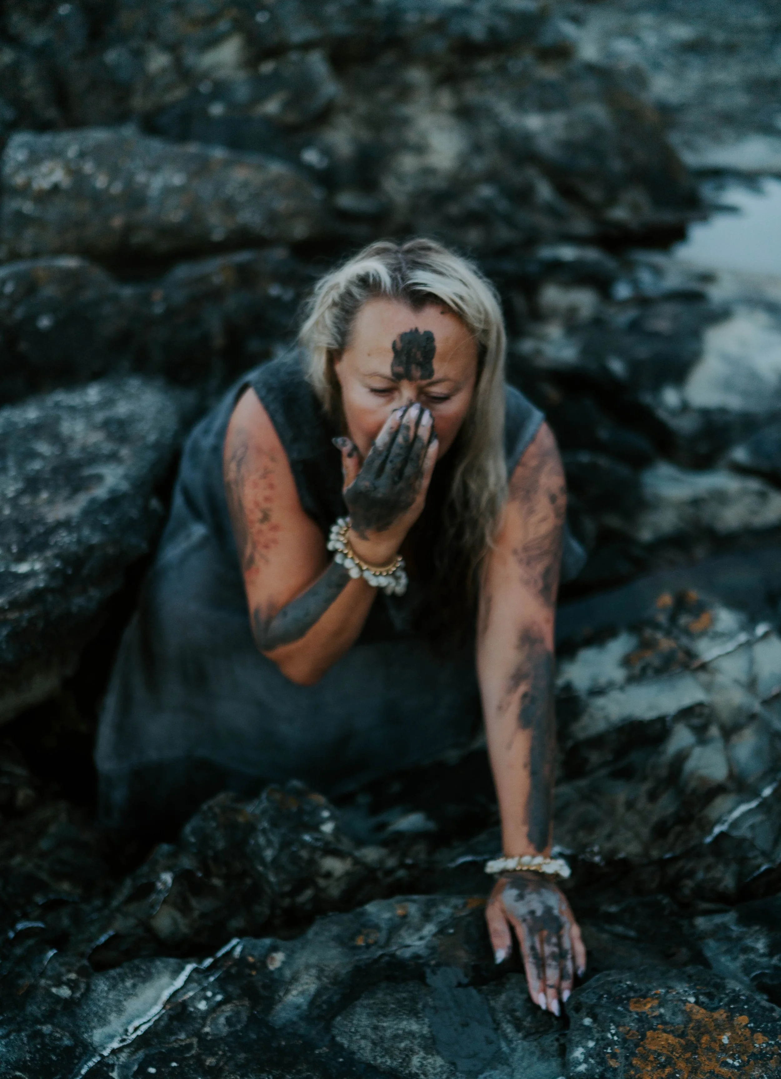 A woman with blonde hair, wearing a sleeveless top and multiple bracelets, is sitting on rocks near water. She appears to be covering her face with her hand, with some mud or dirt on her face, hands, and arms, surrounded by dark rocks.