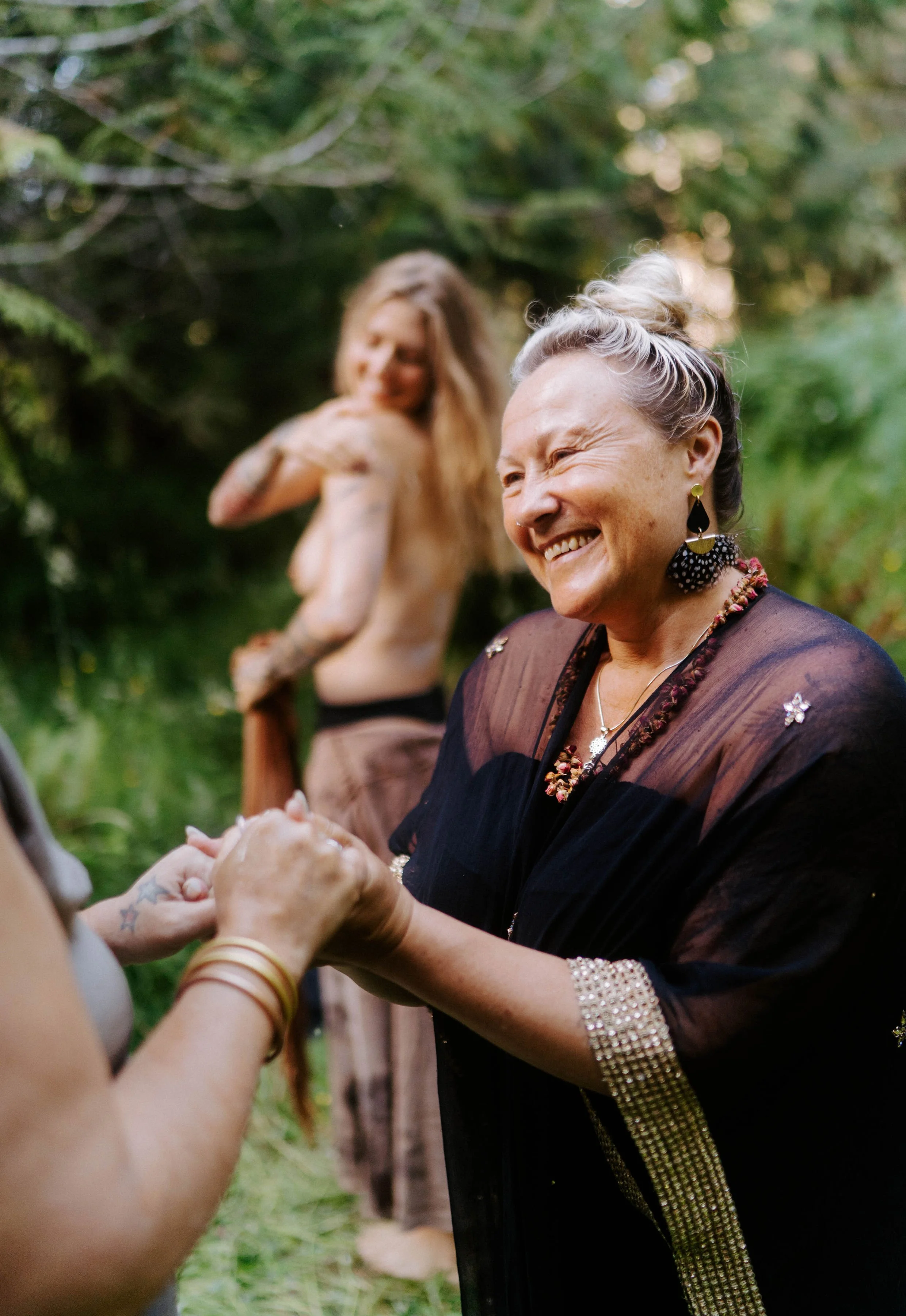 An elderly woman with short gray hair styled in an updo, wearing a black dress with gold embellishments, smiling and holding hands with a person not fully visible. In the background, a woman with long blonde hair and tattoos on her arm, smiling, standing outdoors in a green, wooded area.