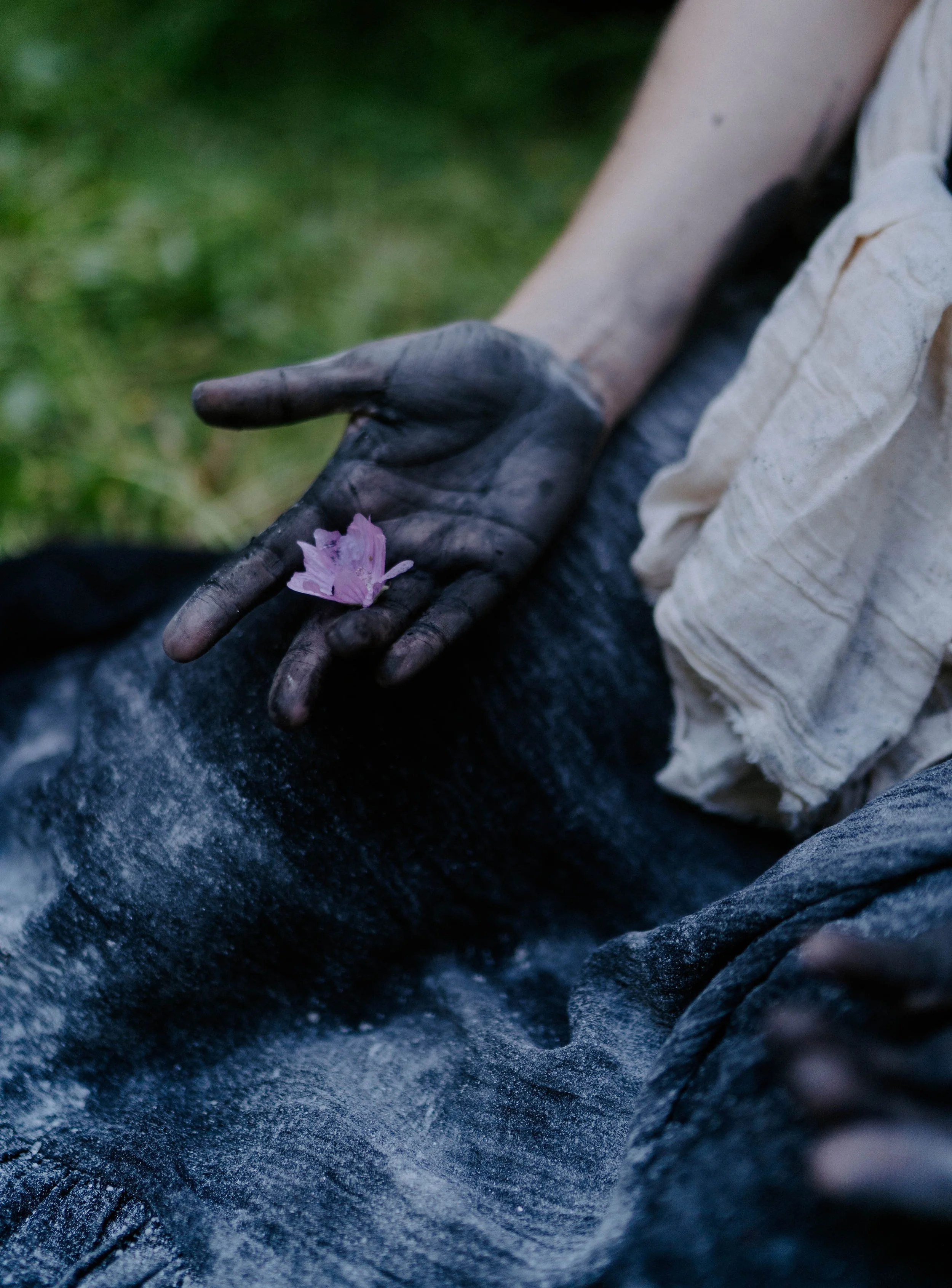 Close-up of a person's hand covered in black soot or dirt, holding a pink cherry blossom flower, with part of their arm and beige clothing visible, set against a green background.