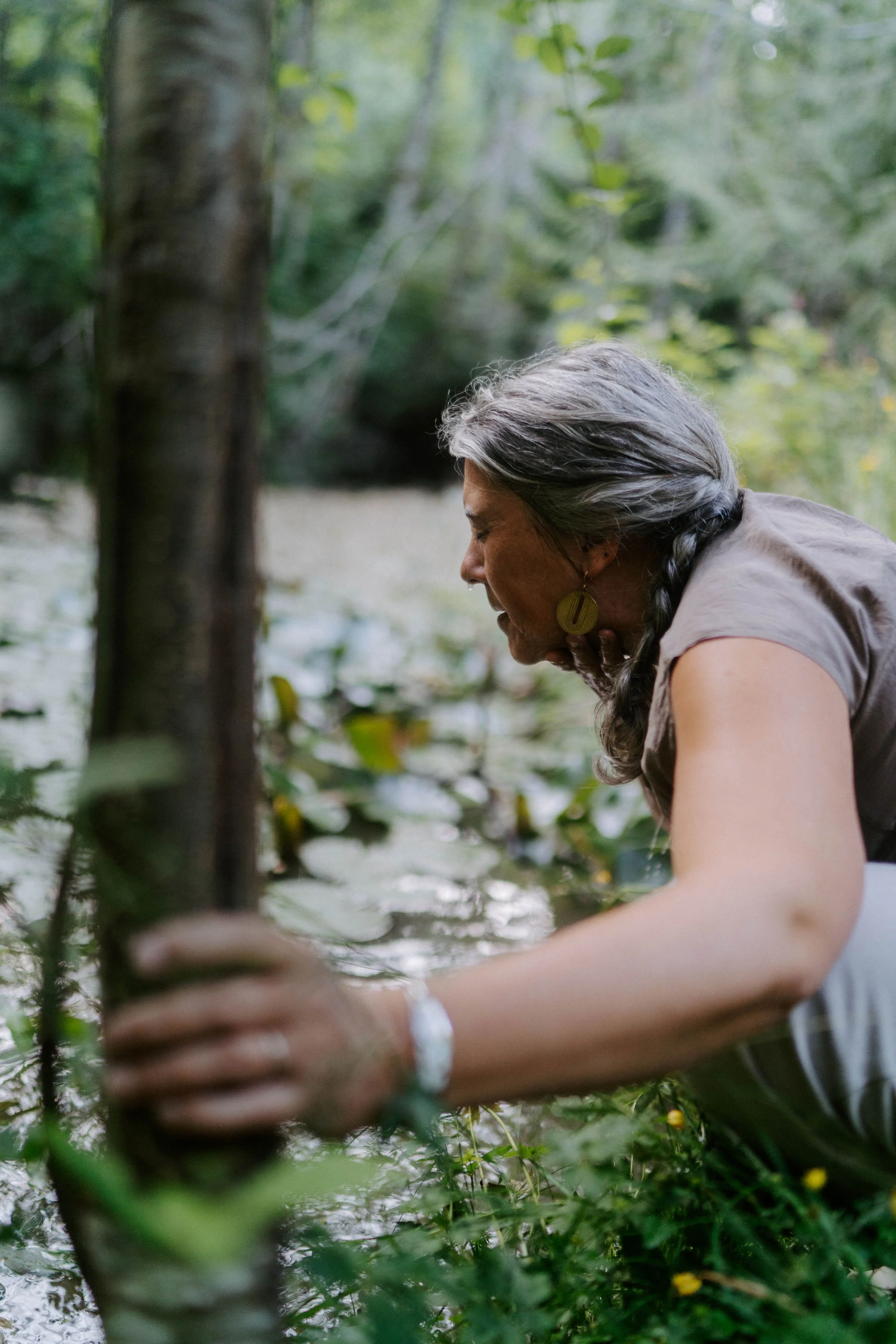 A woman with gray hair braided, wearing a beige top and earrings, leaning over a body of water in a green forest.