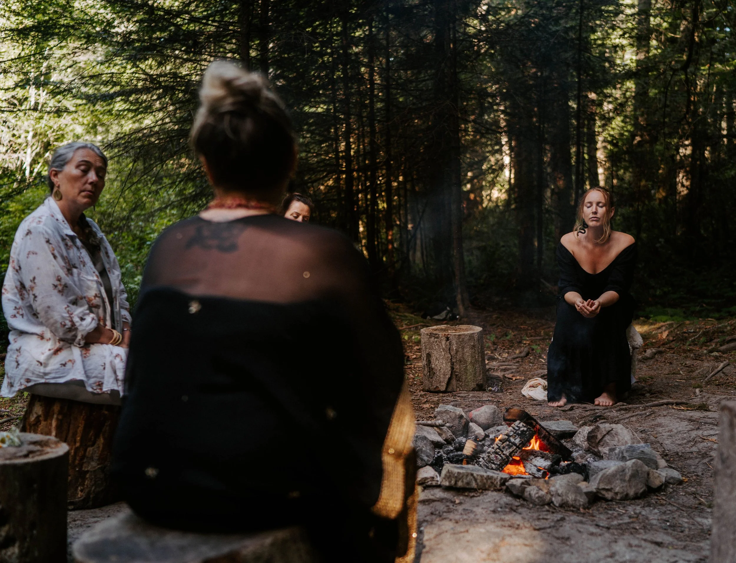 Four women sitting around a campfire in a forest, with sunlight filtering through the trees.