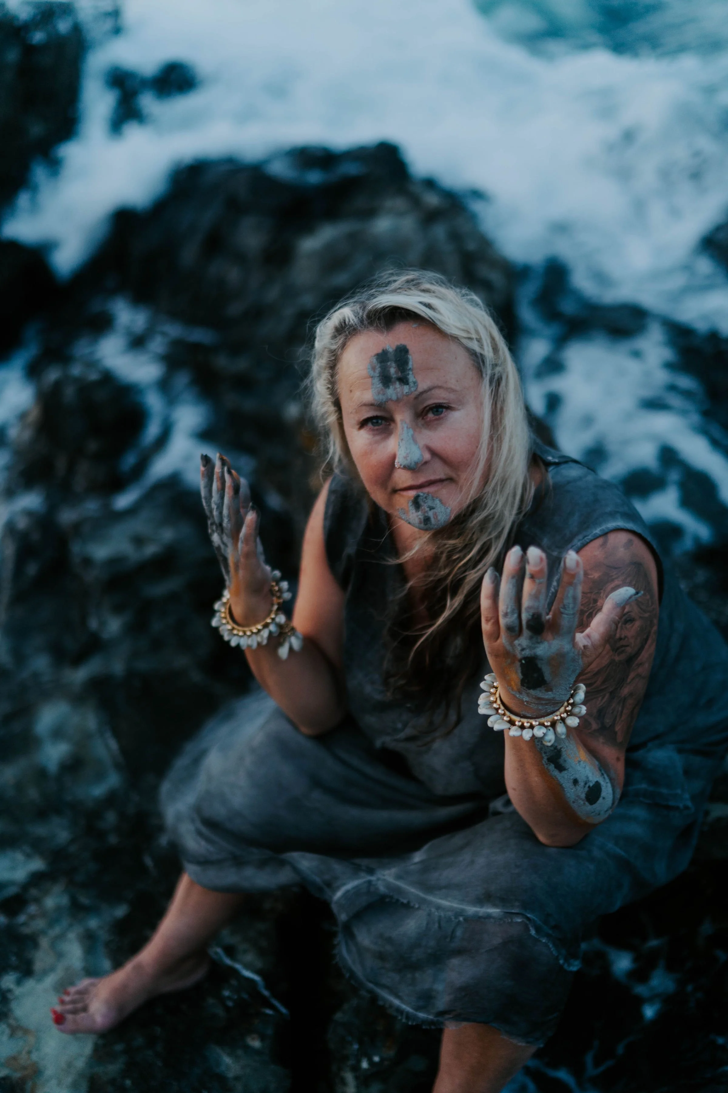 A woman with blonde hair covered in mud, wearing bracelets, at a rocky beach with water and waves in the background.