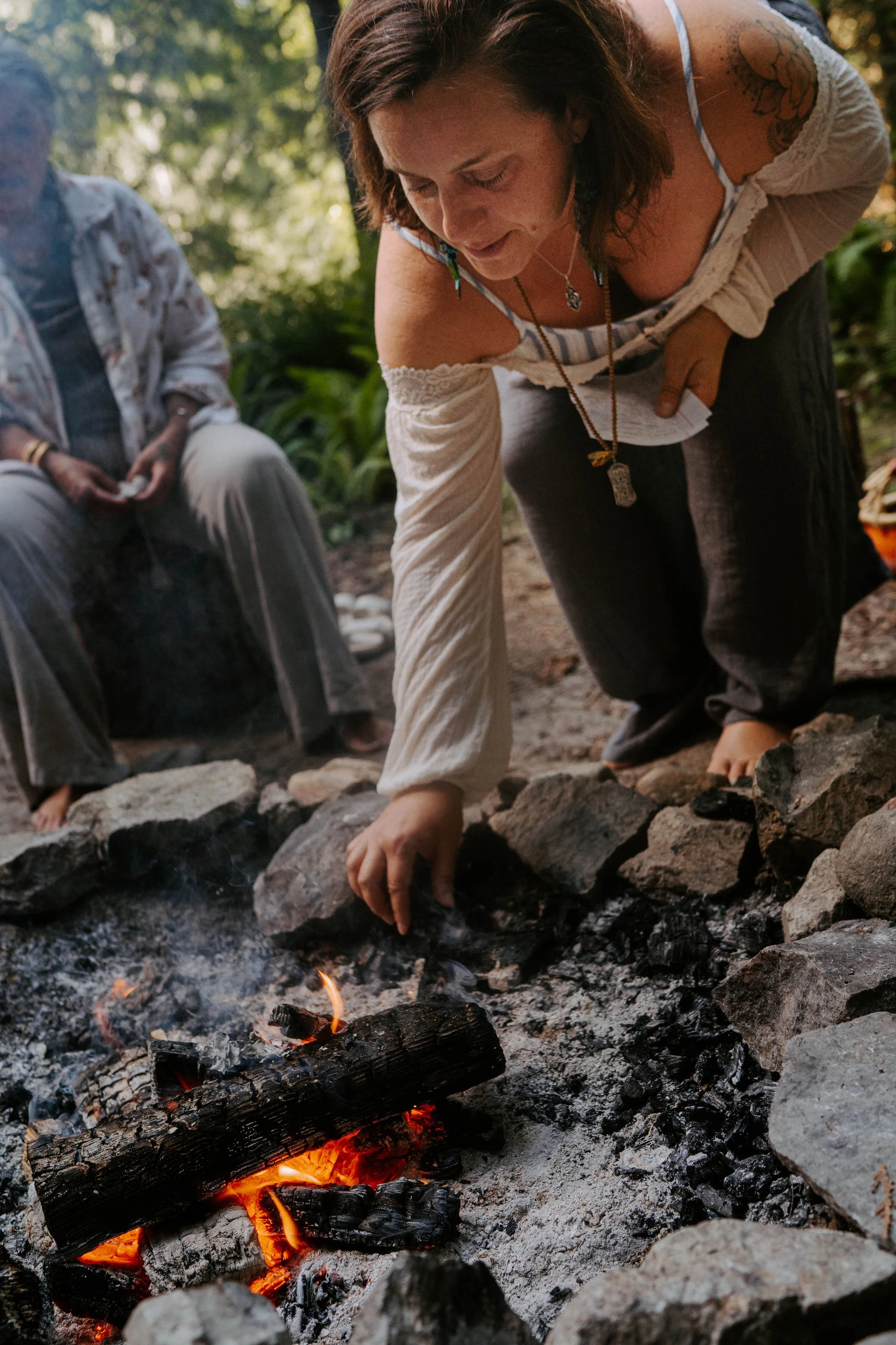Two women are by a campfire in a wooded area; one woman is leaning over the fire, adjusting the logs, while the other woman is sitting nearby. The woman leaning over has short brown hair, tattoos on her arm, and is wearing a light-colored off-shoulder top and necklaces.