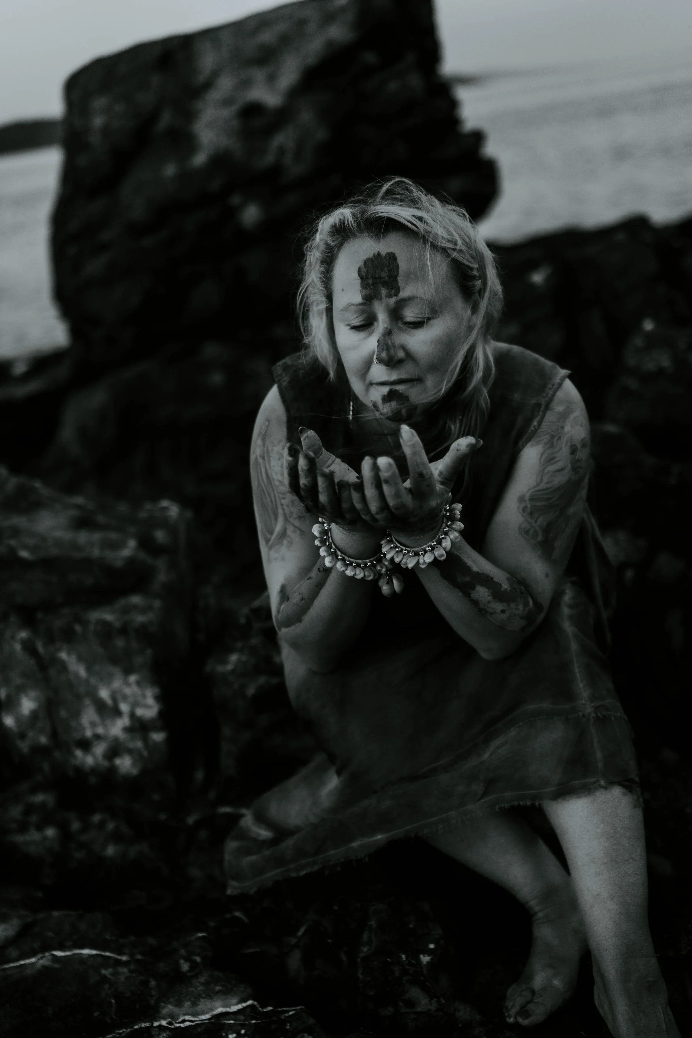 A woman with light skin, tattoos, and jewelry is crouching near rocks by water, holding her hand up to her face with her eyes closed, and there is a large rock formation in the background.