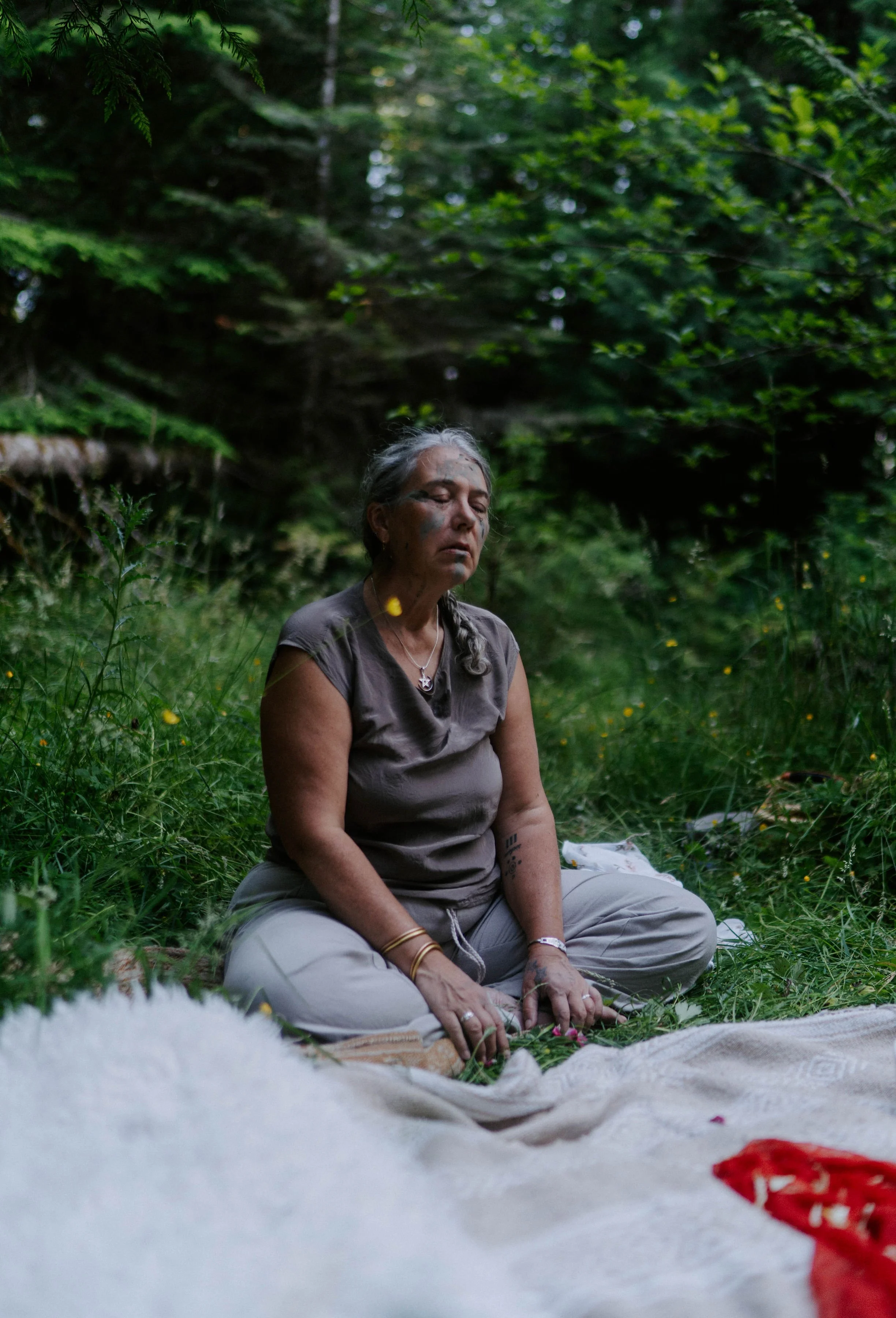 A woman with gray hair sitting cross-legged on the grass, with eyes closed, surrounded by green trees and plants, in a peaceful outdoor setting.