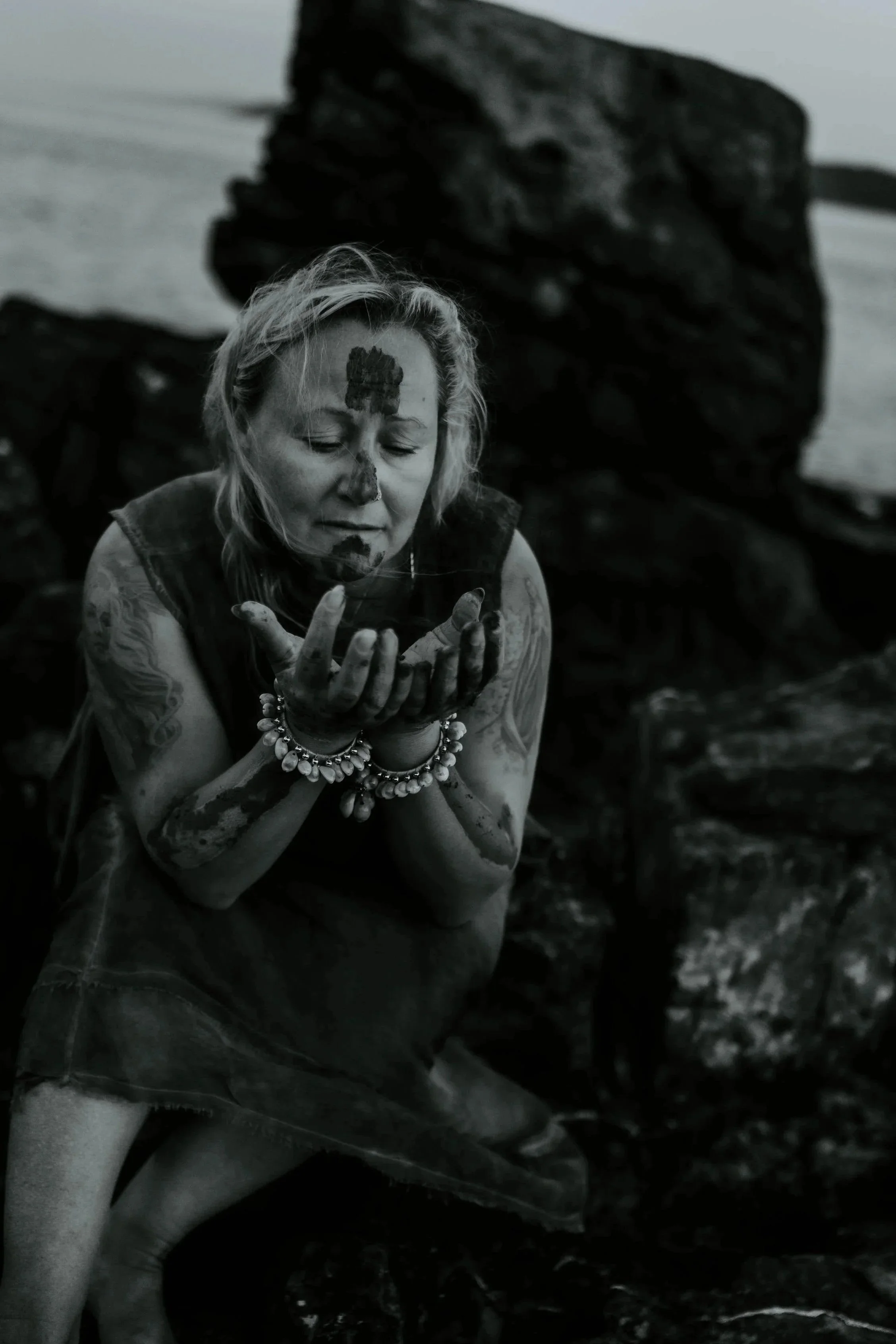 A woman with tattoos and multiple bracelets on her wrists, eyes closed, holding her hands together near her face, with dark rocks and water in the background.