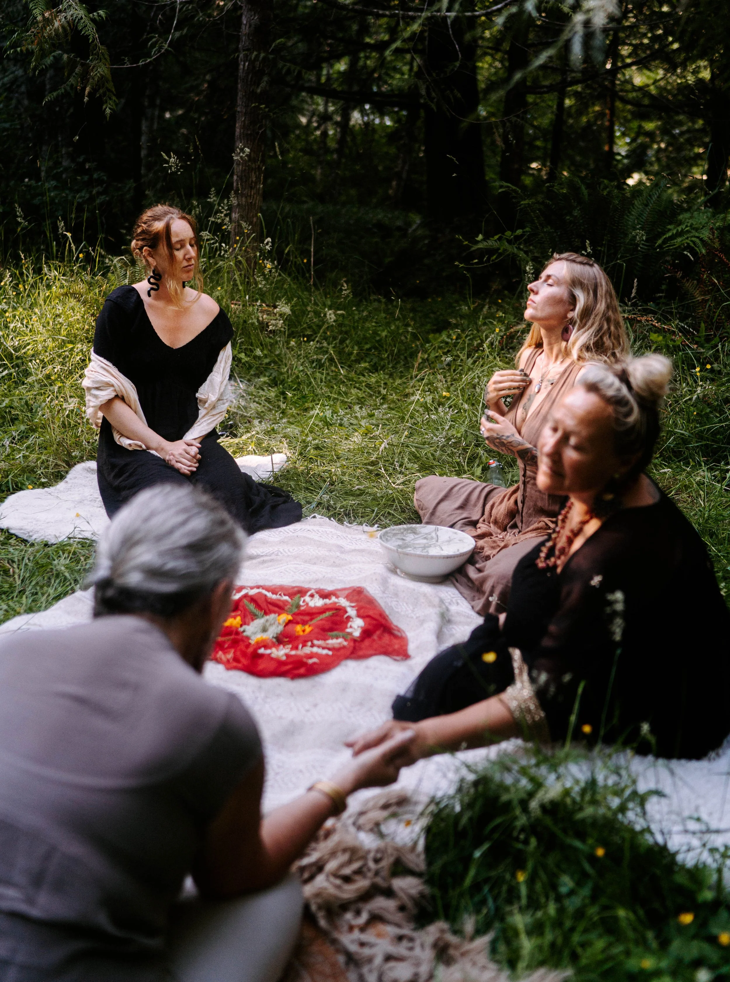 Group of women gathered outdoors in a forest, sitting on a blanket with flowers, participating in a spiritual or meditative ceremony.