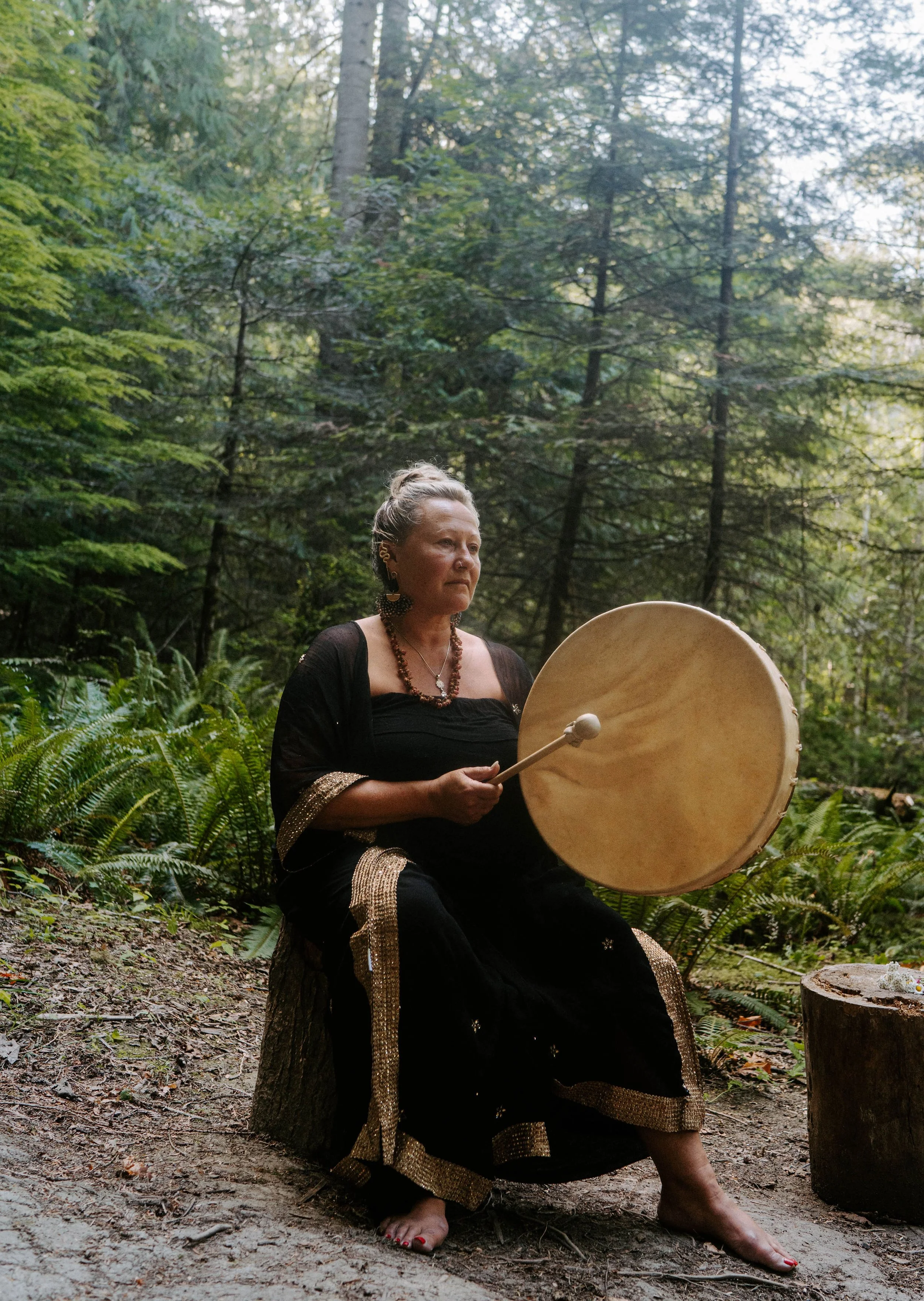 A mature woman with silver hair, dressed in a black and gold-embellished gown, seated on a tree stump in a forest, holding a large drum and a mallet, surrounded by lush green foliage and tall trees.
