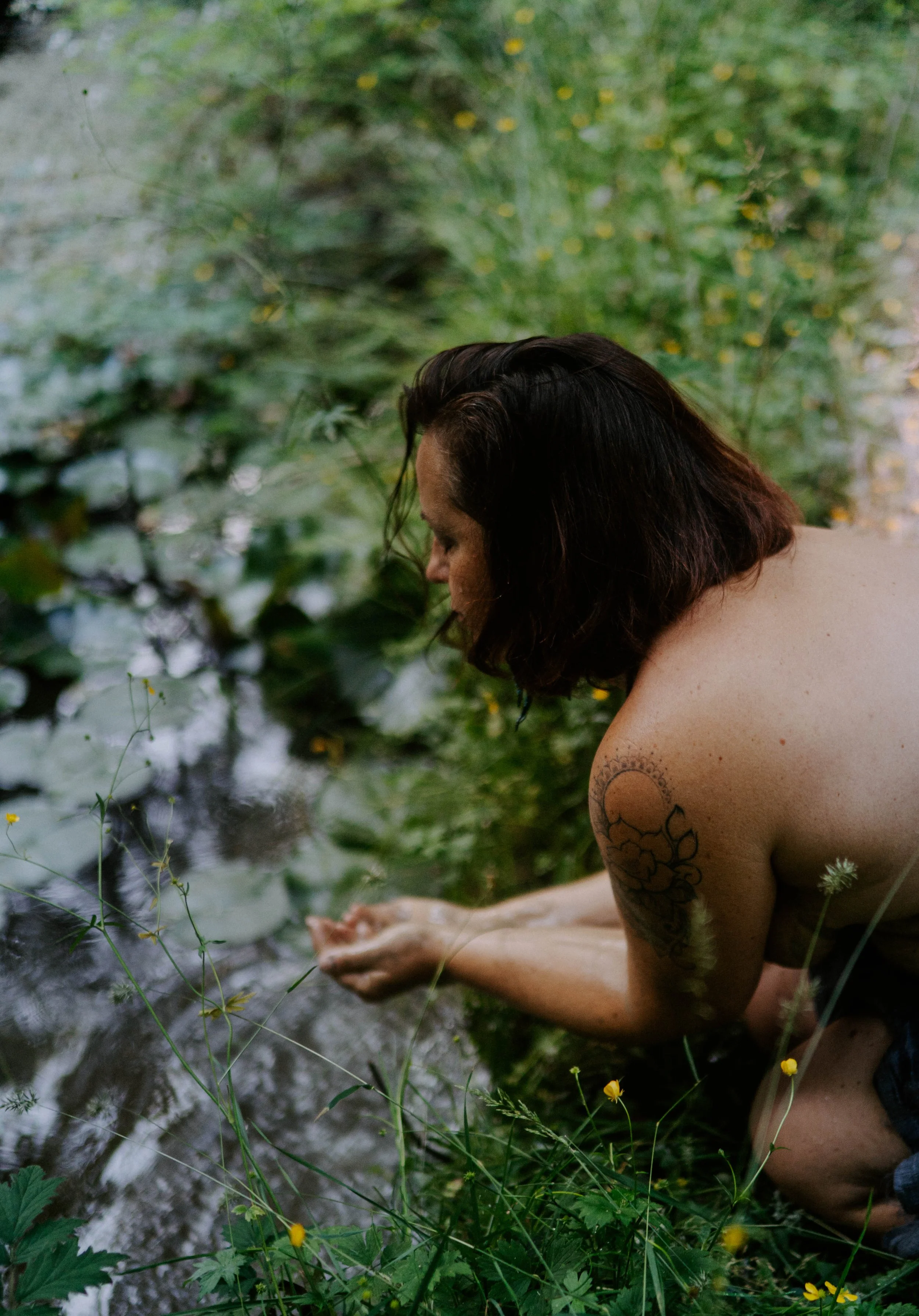 A woman with tattoos on her arm sitting by a creek, looking down at the water with her hands clasped, surrounded by green plants and yellow flowers.