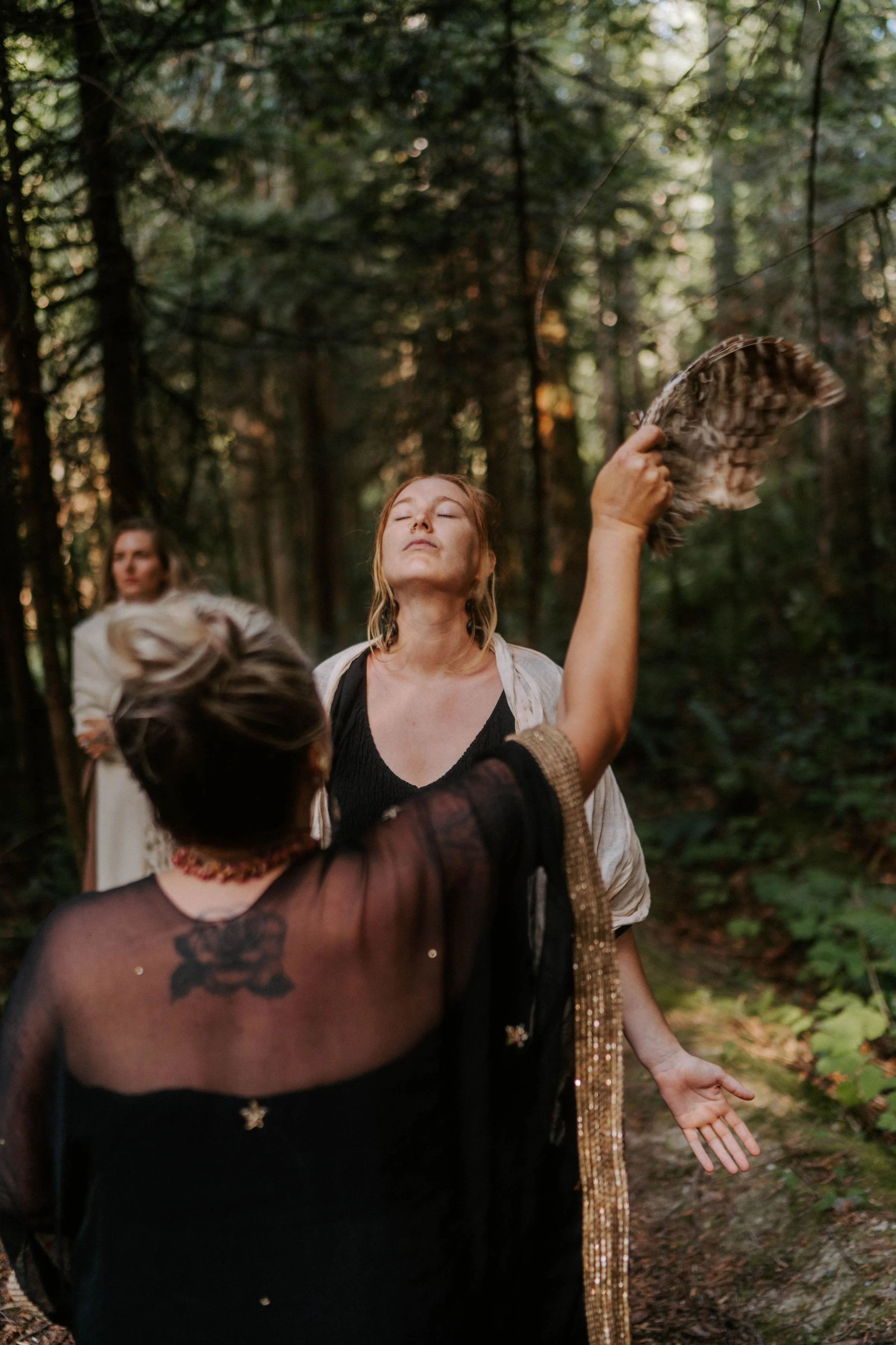 People in an outdoor forest setting, with one woman holding a bird feather and another person possibly touching her shoulder.