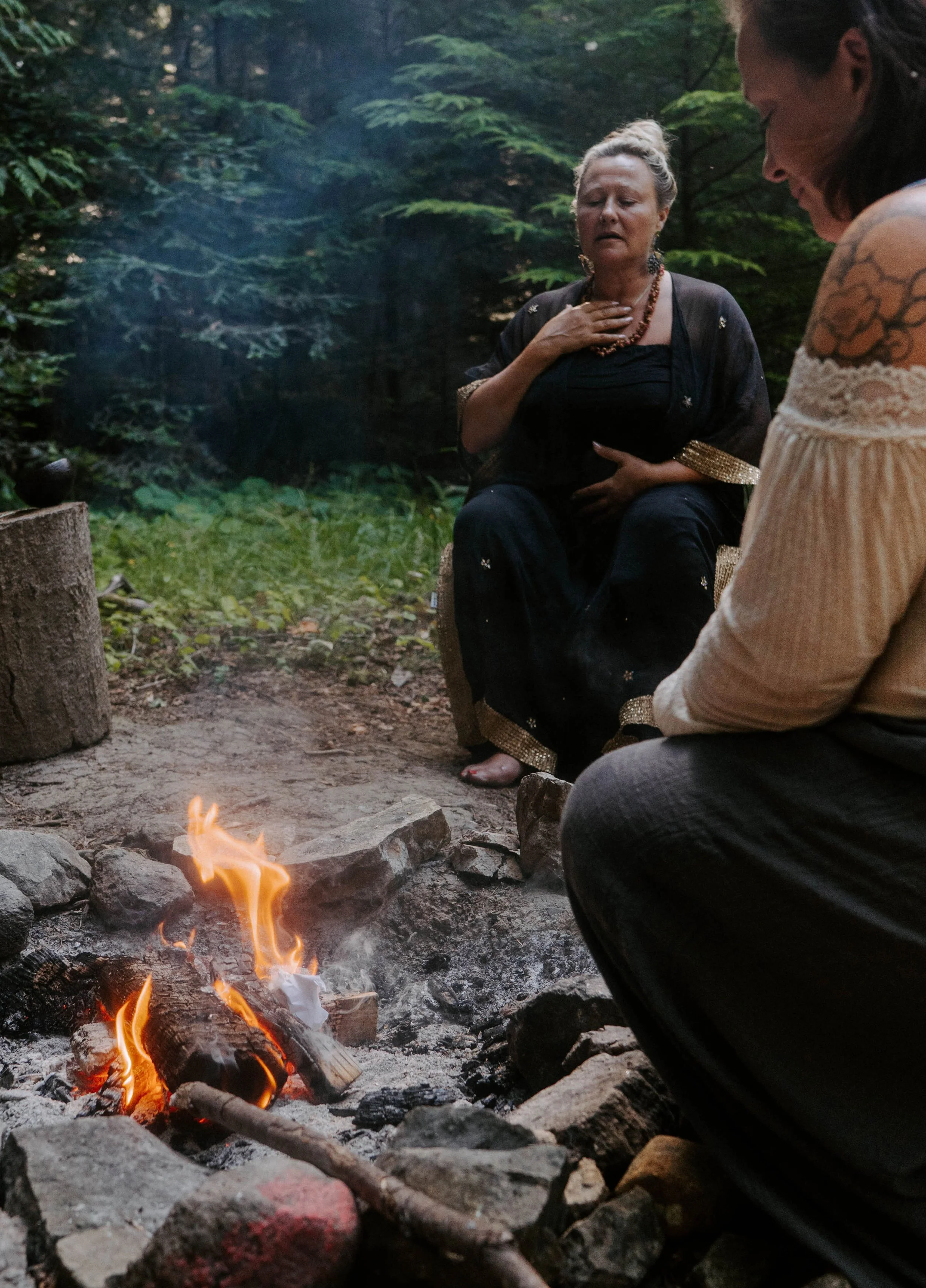 Two women sit around a campfire in a forest, one with hand on her chest and the other with head bowed, in a solemn moment.
