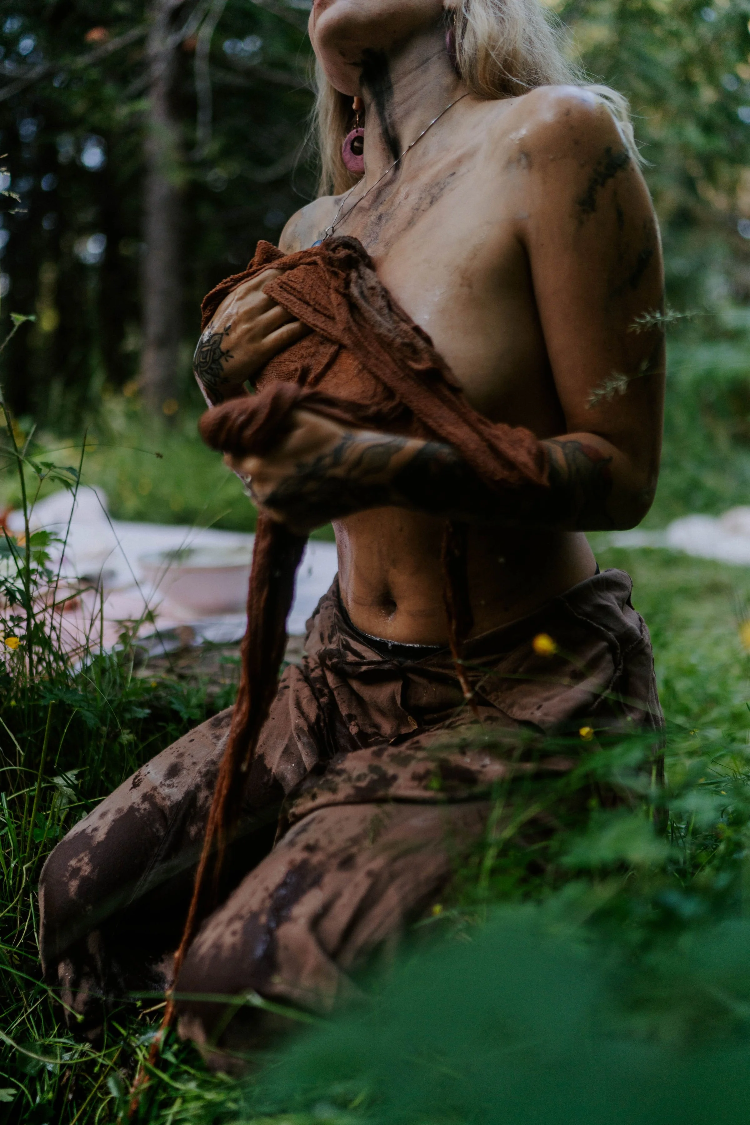 A person with tattoos and piercings, wearing camouflage pants, is shirtless and holding a brown towel or piece of cloth. They are outdoors in a forest or wooded area, with greenery and trees in the background.