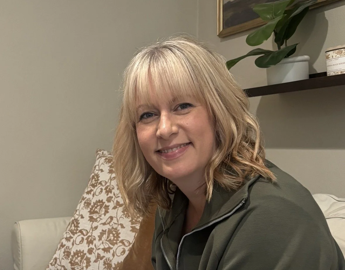 A smiling woman with blonde hair seated on a sofa, with a decorative pillow and a shelf with plants and decor in the background.