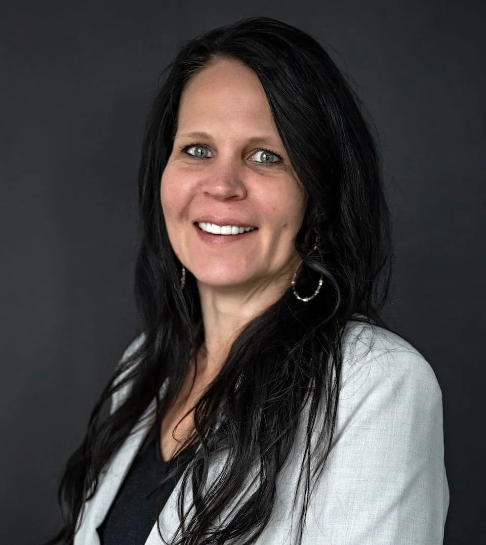 A woman with long black hair, wearing a white blazer over a black top, and hoop earrings, smiling against a dark background.