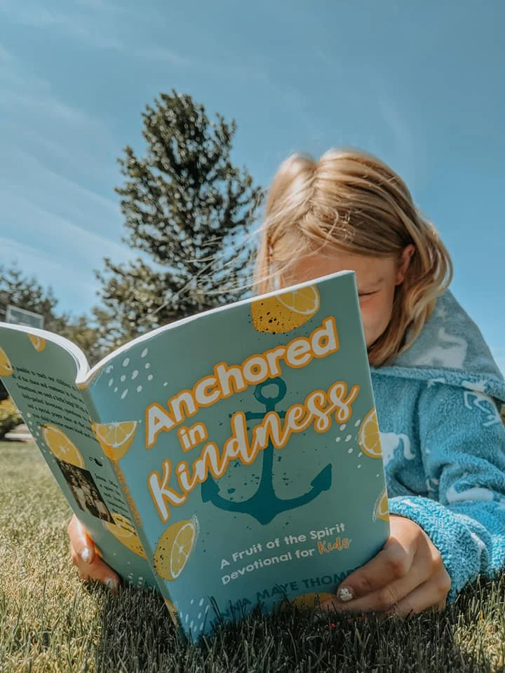 A young girl reading a book titled 'Anchored in Kindness' outdoors on grass with a tree and blue sky in the background.