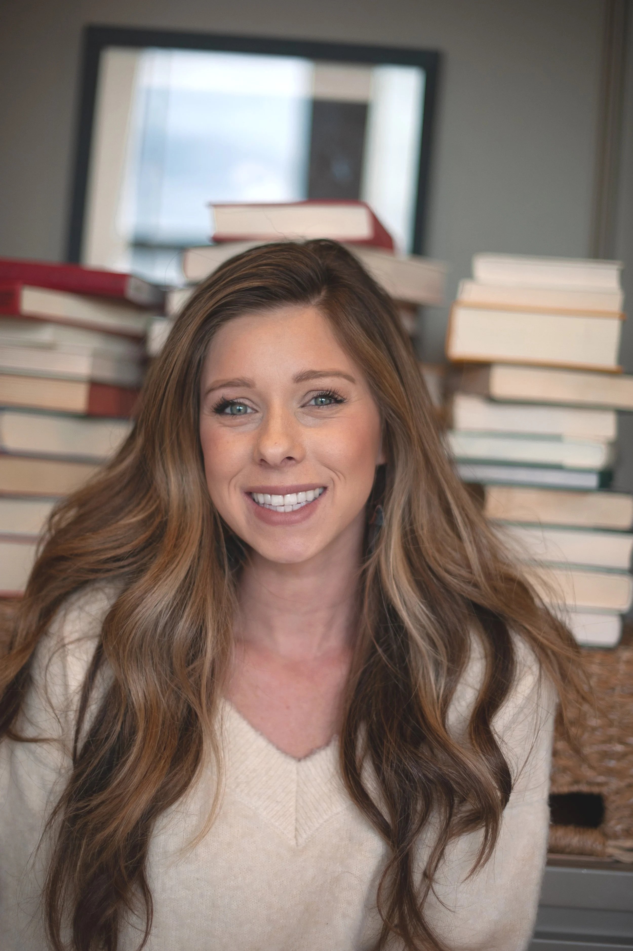 A woman with long, wavy brown hair smiling in front of a stack of books in an indoor setting.