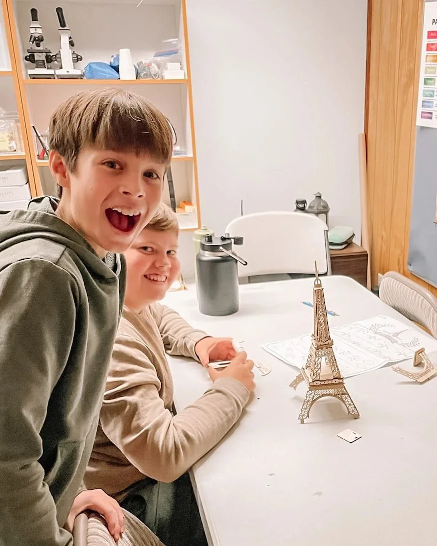 Two boys smiling at a table with a miniature Eiffel Tower model, in what appears to be a classroom or science lab setting.