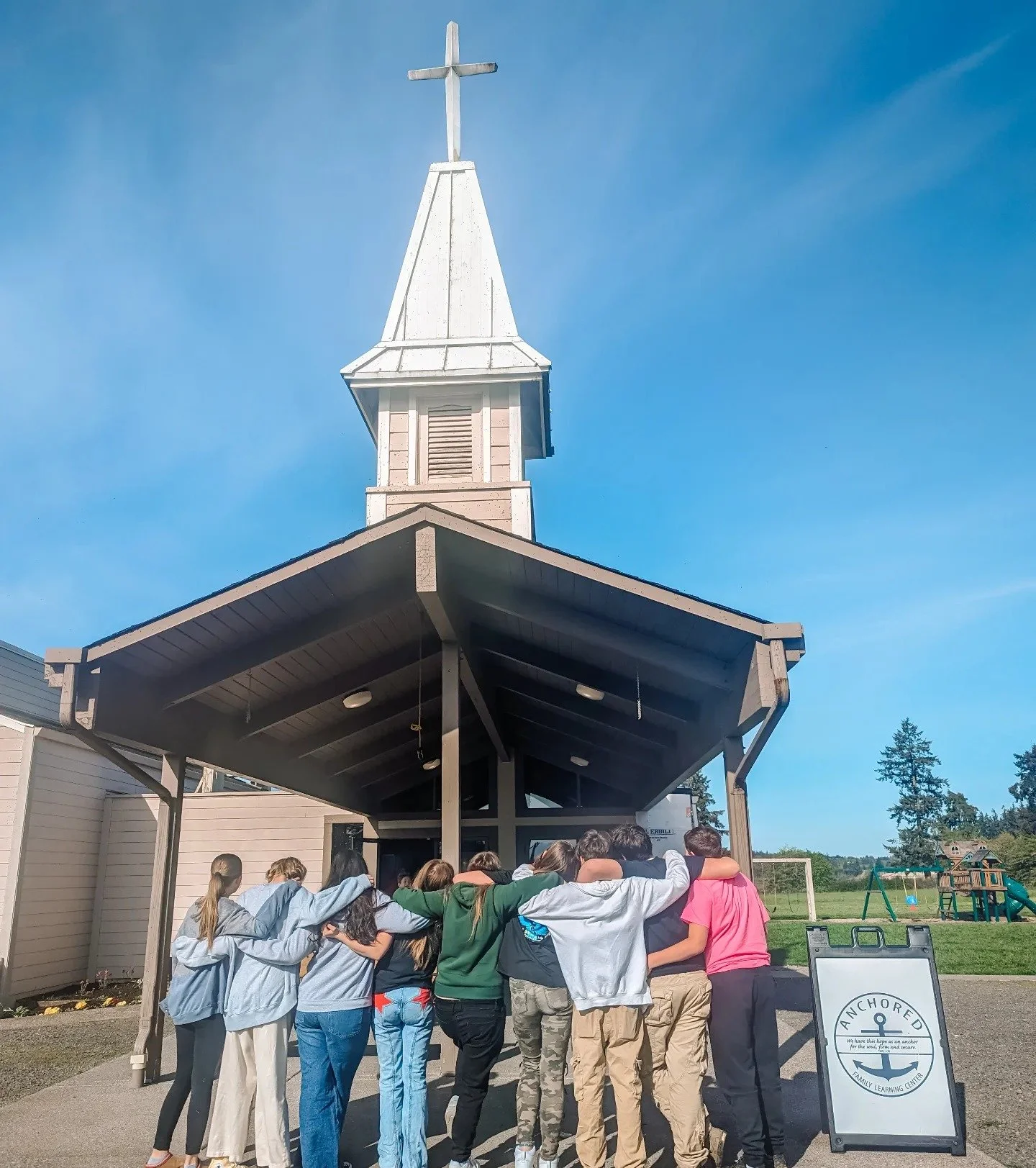A group of people standing in a huddle under a covered outdoor structure with a church steeple and cross on top, located at a family learning center named 'Anchored.' School play equipment is visible in the background.