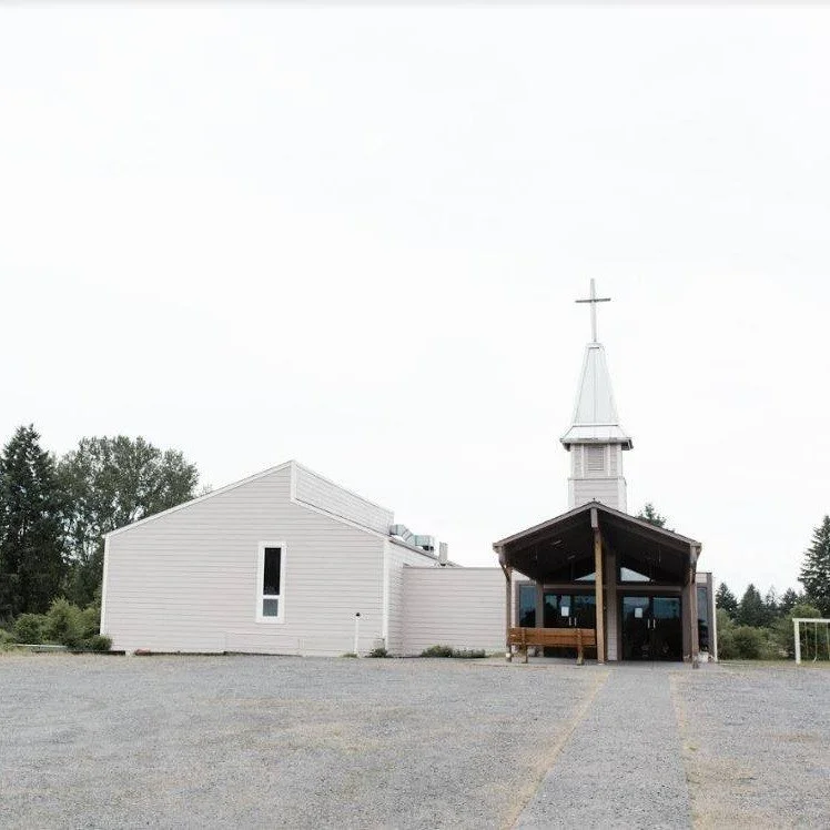 White church with a steeple and cross, and an adjacent building, on a gravel lot with trees in the background.