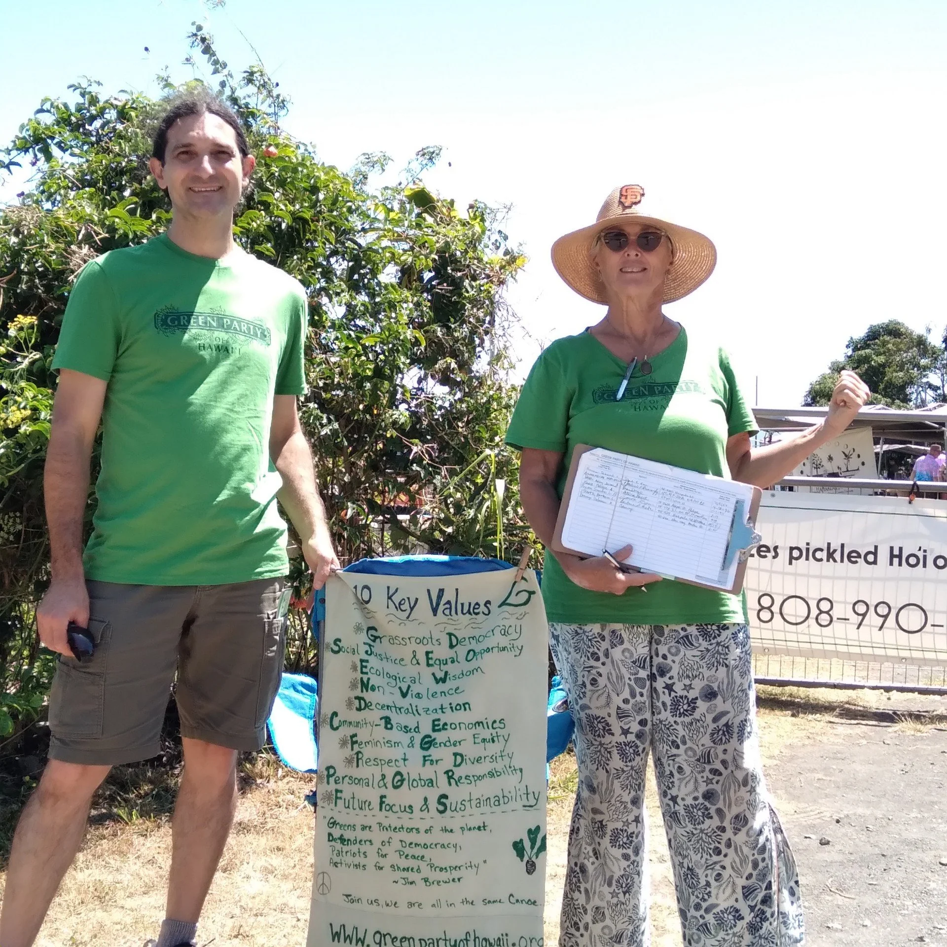 Two people standing outdoors at a rally or event with a large sign that reads '10 Key Values' with green and black text about social justice, democracy, eco-friendliness, diversity, and sustainability. The person on the left is a man with short dark hair wearing a green T-shirt and khaki shorts. The person on the right is a woman with blonde hair, sunglasses, a wide-brimmed straw hat with an SF logo, a green T-shirt, and patterned pants, holding a clipboard and a smaller sign.