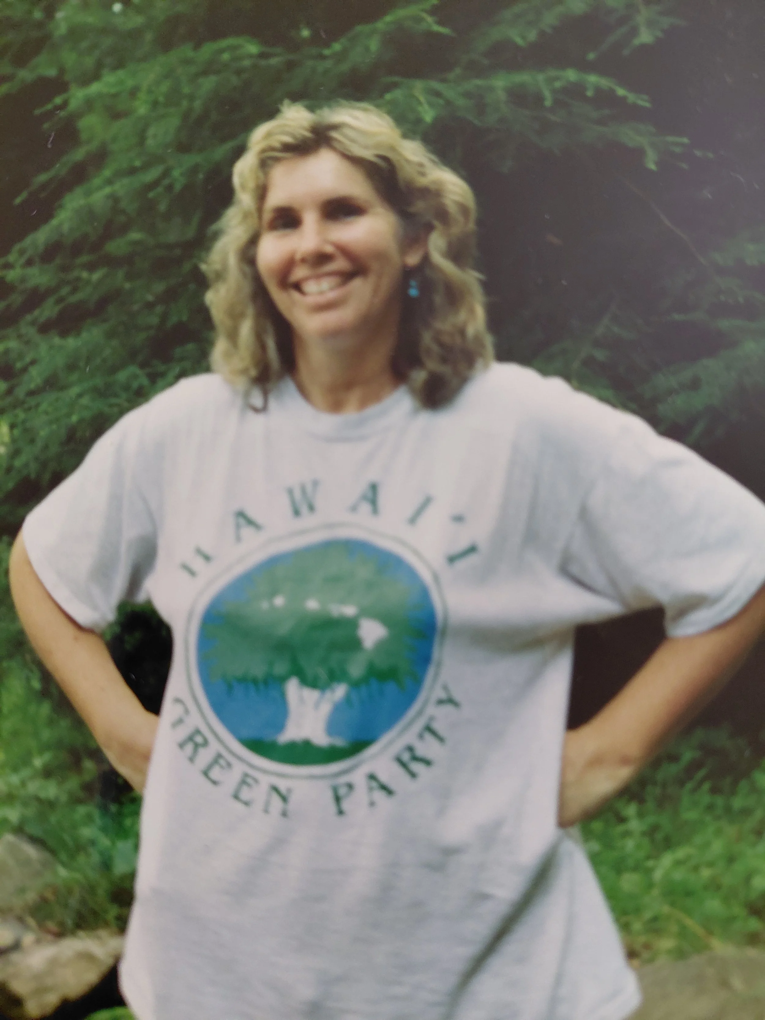 A woman with blonde, curly hair smiling outdoors in front of green trees, wearing a white T-shirt with a circular logo that says "Evergreen Park".