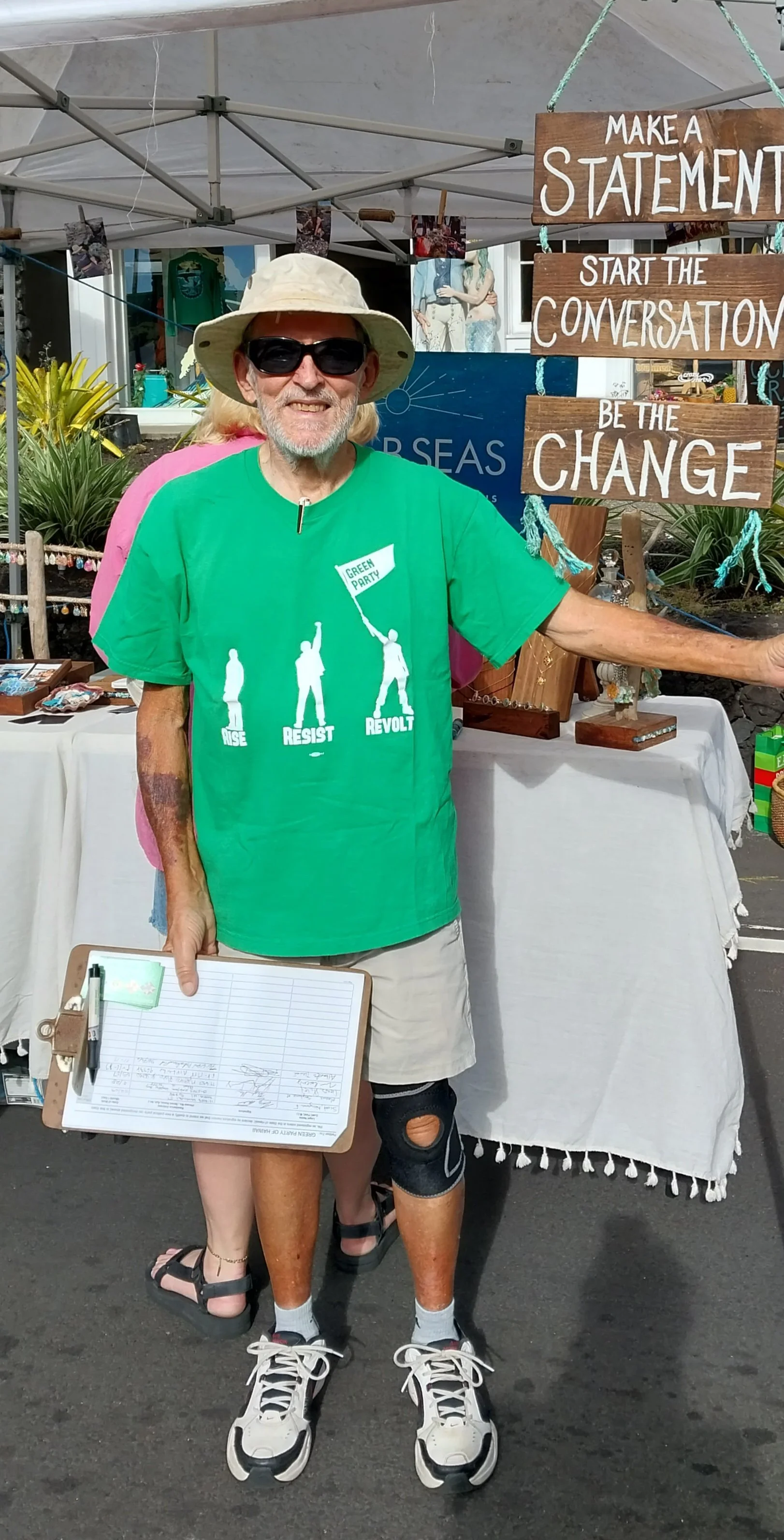 A man wearing a green T-shirt, beige shorts, a beige sun hat, sunglasses, and sneakers, standing at an outdoor event booth holding a clipboard. Behind him are signs that read: "Make a Statement," "Start the Conversation," and "Be the Change." The booth has various items and plants visible.