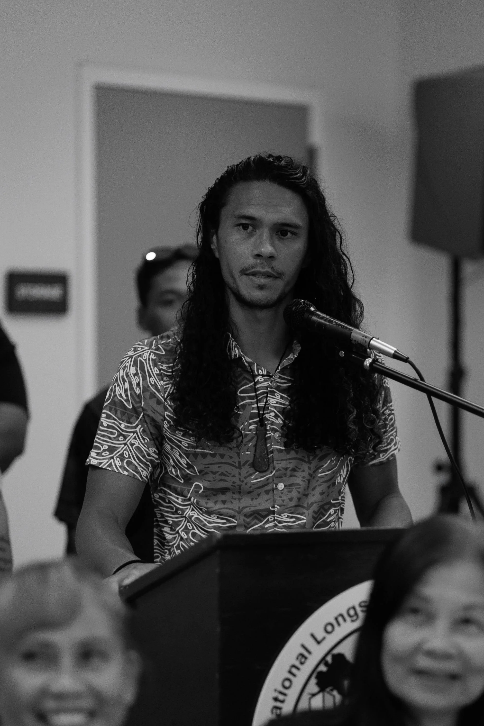 A man with long curly hair speaking into a microphone during an event, with people in the background and a logo partially visible in the foreground.