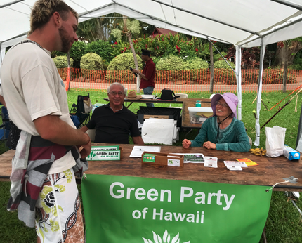 People at a Green Party of Hawaii booth outdoors under a tent, with a man talking to two seated women.