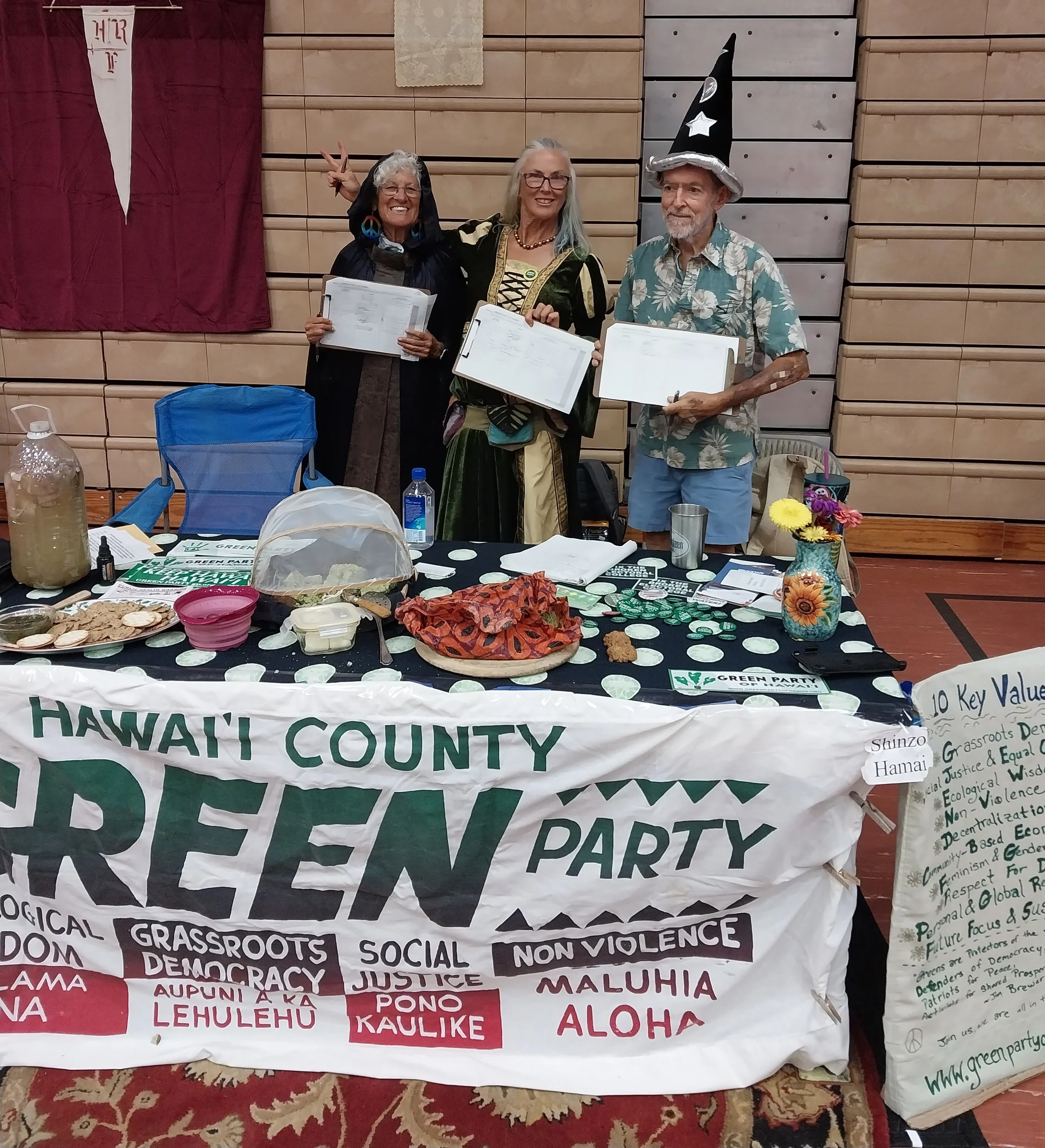 People at an event holding documents, with a table filled with food, drinks, and informational materials related to a Hawaiian-themed or environmental cause, and a banner supporting a green party or social justice movement in Hawaii.