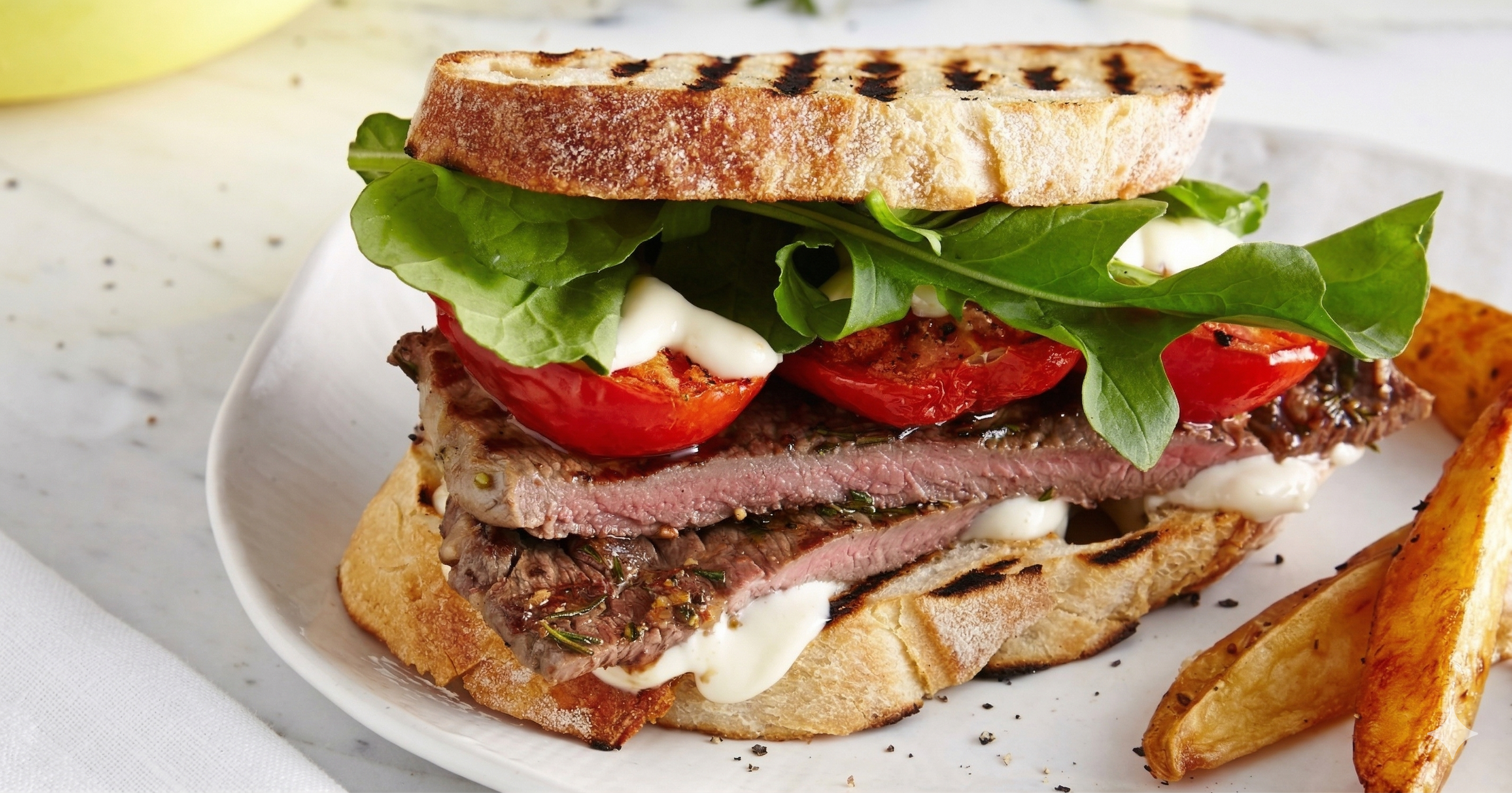 A close-up of a steak sandwich with grilled bread, fresh greens, tomato slices, and white sauce, served with potato wedges.